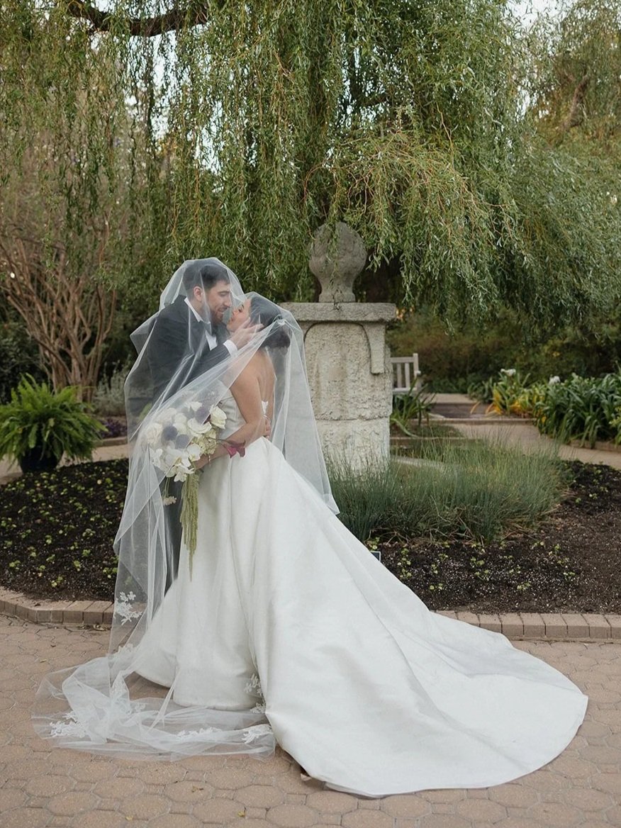 A newlywed couple sharing a kiss, with the bride holding a bouquet, standing under a tree with hanging branches in a garden setting.