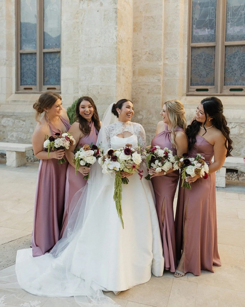 A bride and five bridesmaids standing outside in front of a stone building, all holding bouquets and smiling at each other.