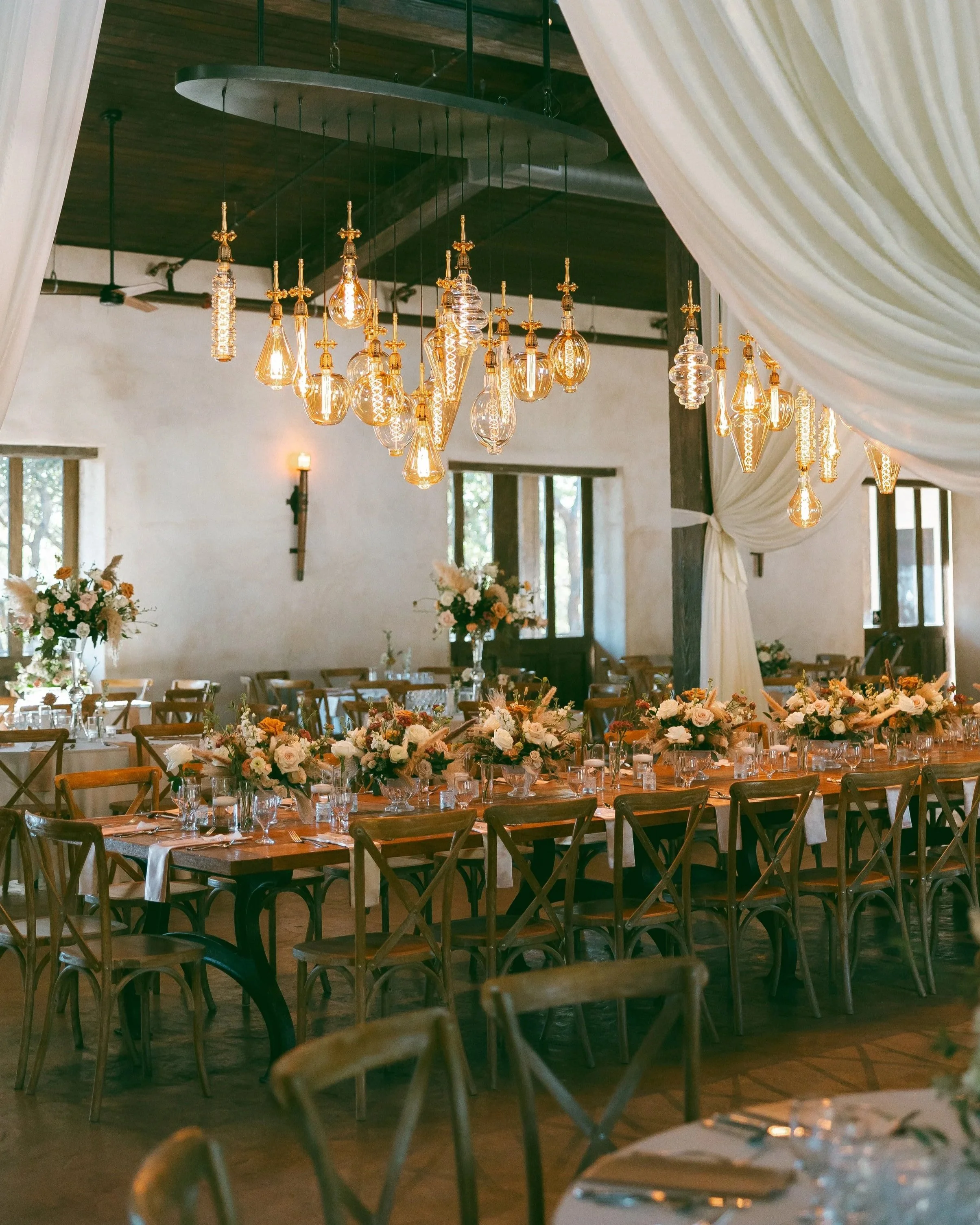 Elegant indoor wedding reception with wooden tables decorated with floral centerpieces, surrounded by wooden chairs. A modern chandelier with hanging glass bulbs and draped curtains enhance the rustic yet sophisticated ambiance.