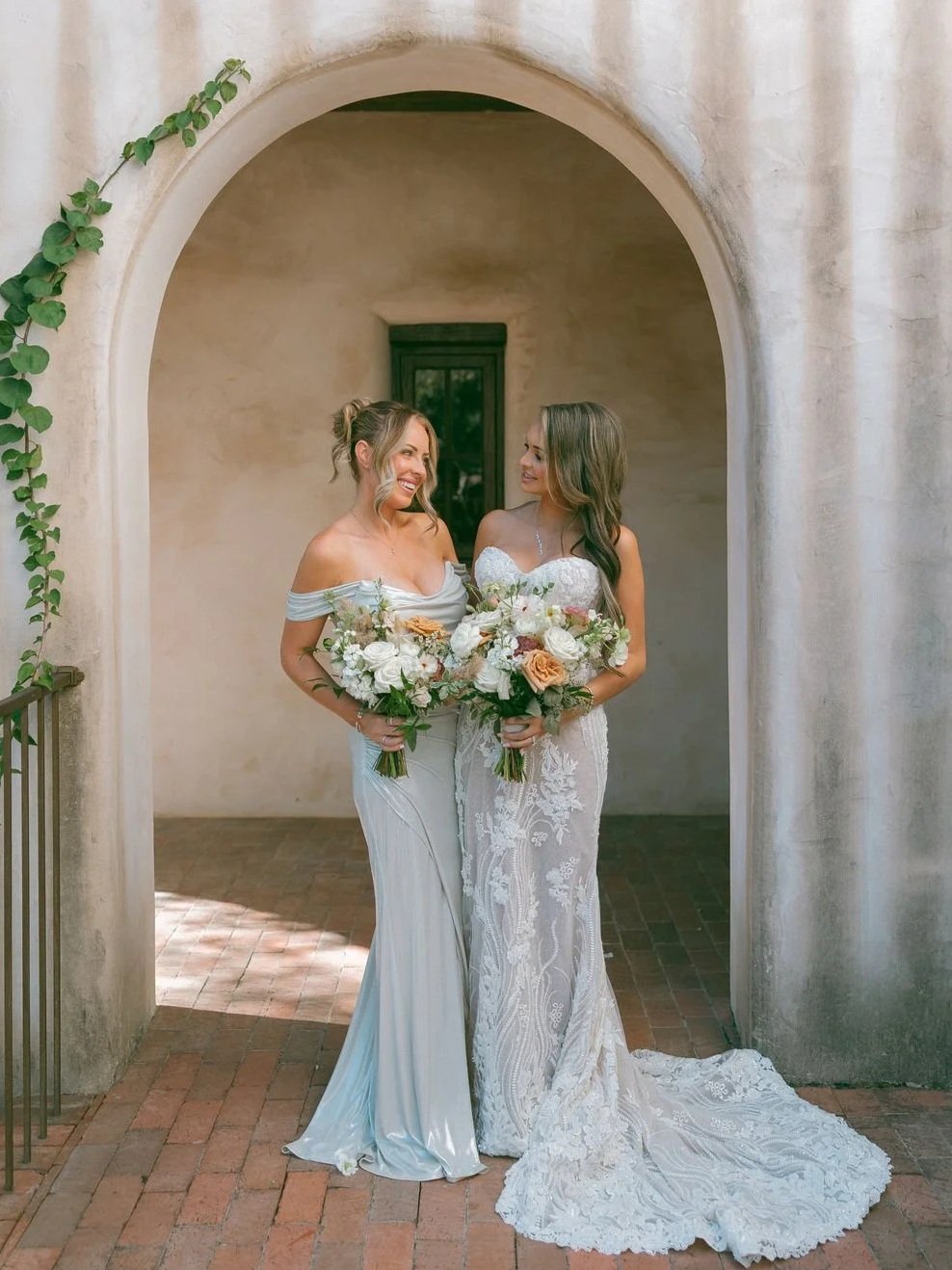 Two women in wedding dresses holding bouquets of flowers, standing under an archway with a beige wall background.