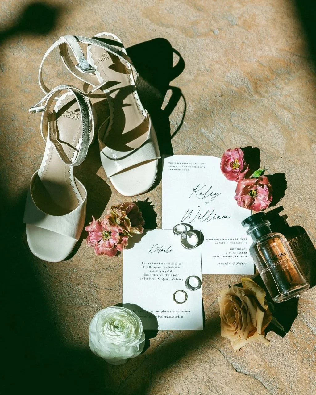 Flat lay of wedding items including white high-heeled sandals, pink and cream flowers, wedding invitation with names Kaley and William, rings, a bottle of perfume, and a rose on a textured surface with sunlight.