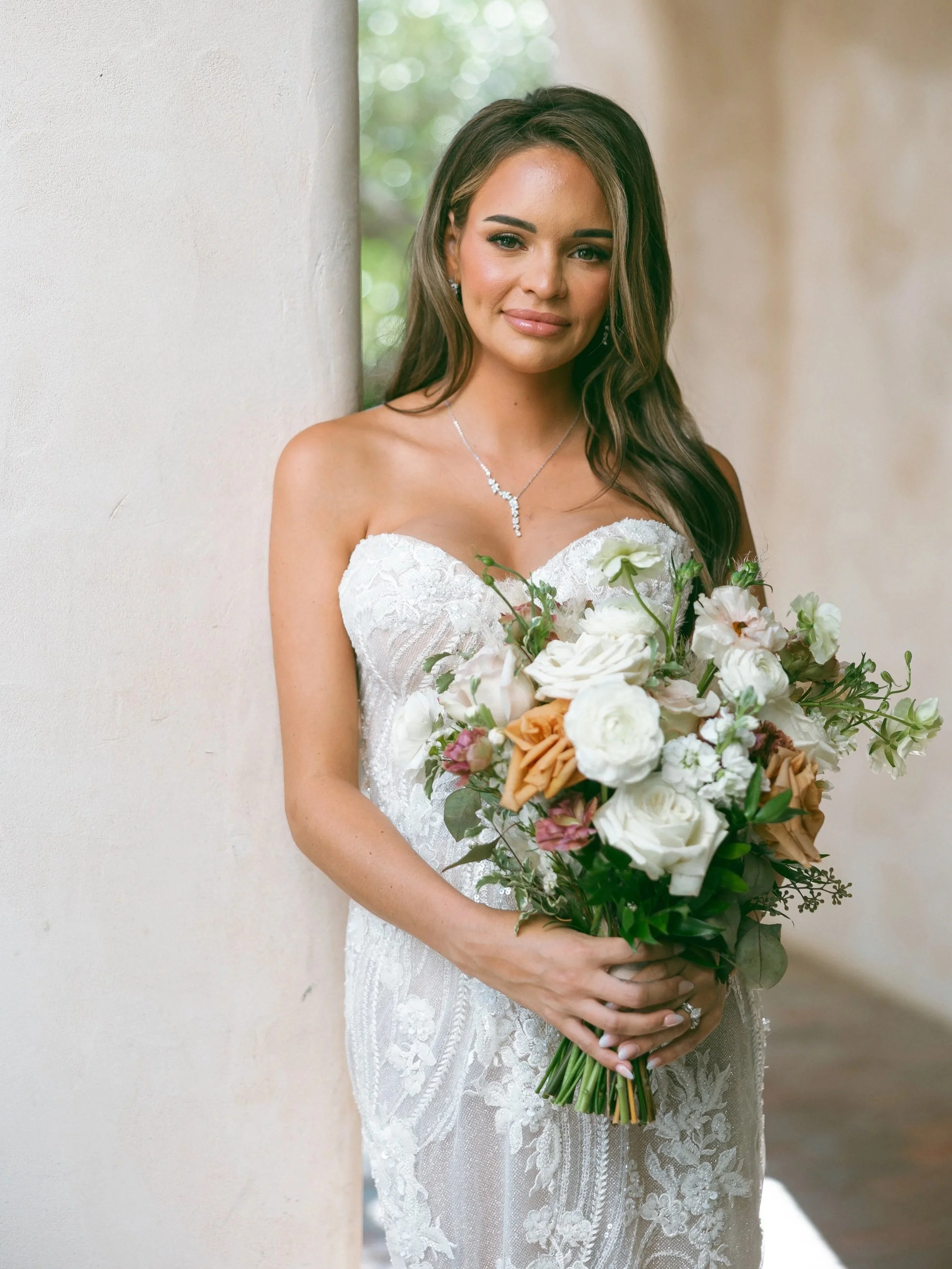 A bride in a strapless white wedding dress holding a bouquet of white, peach, and pink flowers, standing indoors by a beige wall, looking at the camera.