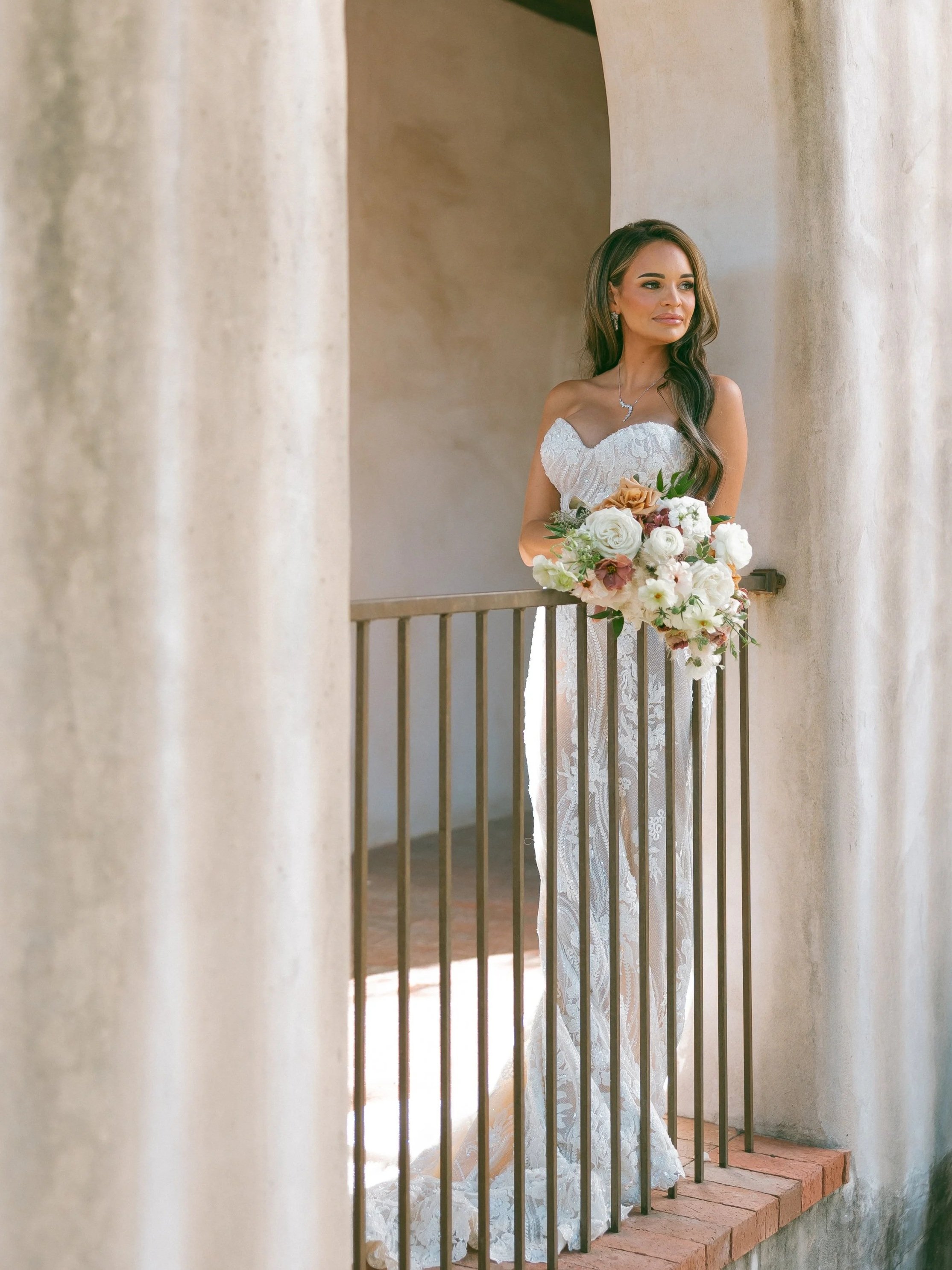 A bride wearing a white off-the-shoulder lace wedding dress, holding a bouquet of white and blush flowers, standing on a small balcony with brick flooring, framed by beige stone columns.