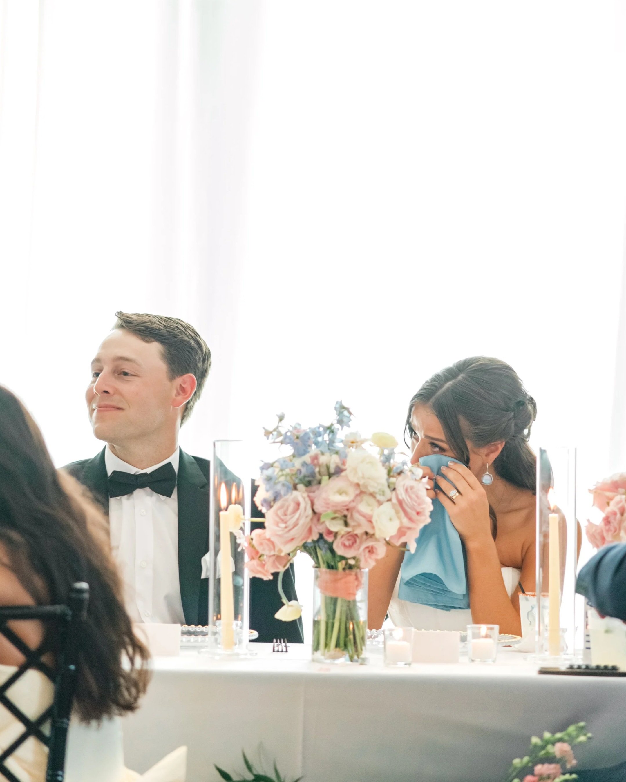 A woman at a wedding reception sitting at a table with a man. The woman appears emotional, holding a tissue or napkin to her face. The man is dressed in a tuxedo with a bow tie, smiling softly. The table is decorated with pink and white flowers, cand