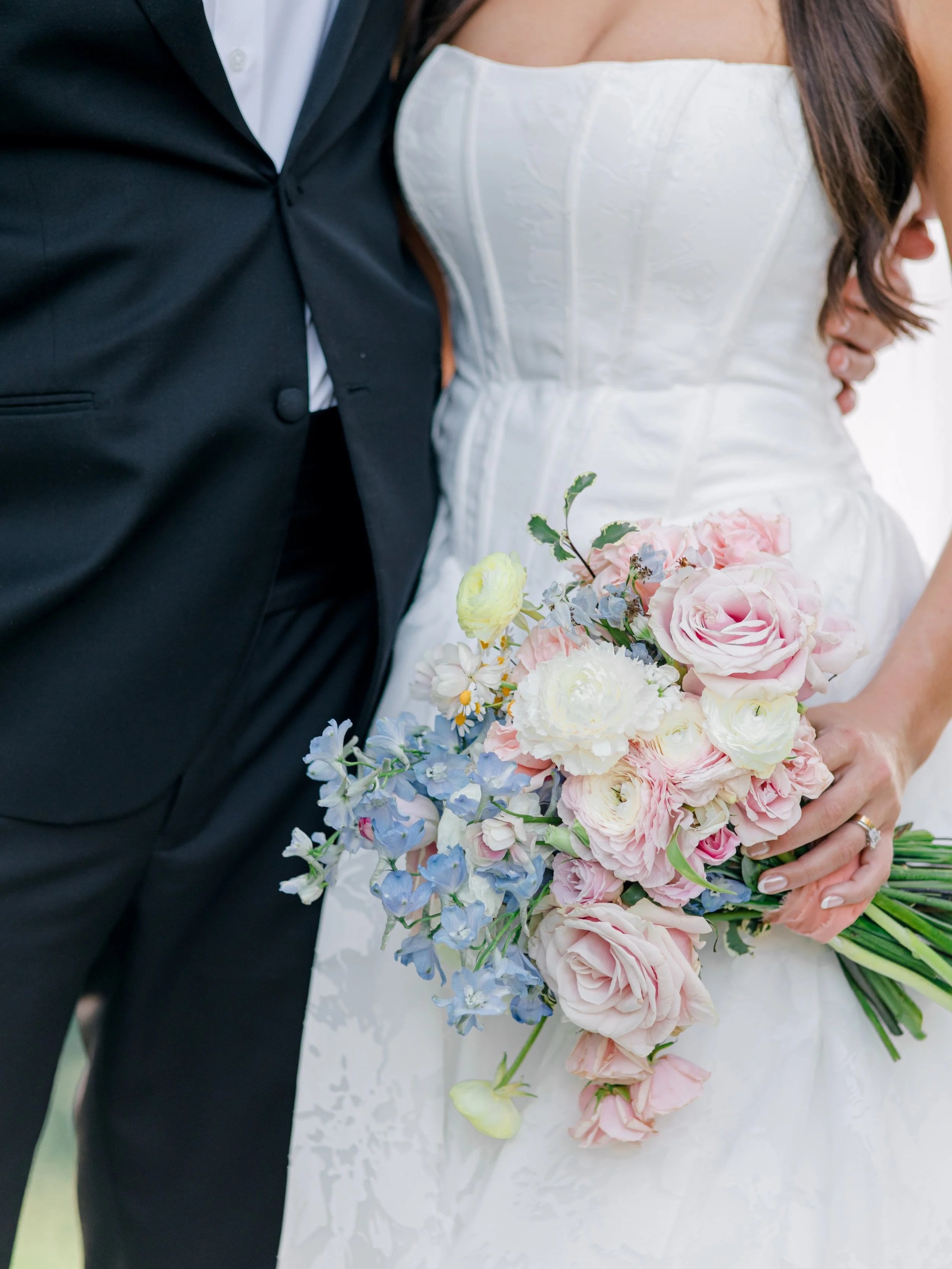 Close-up of a bride holding a bouquet of pink, white, and blue flowers, standing next to a groom dressed in a dark suit.
