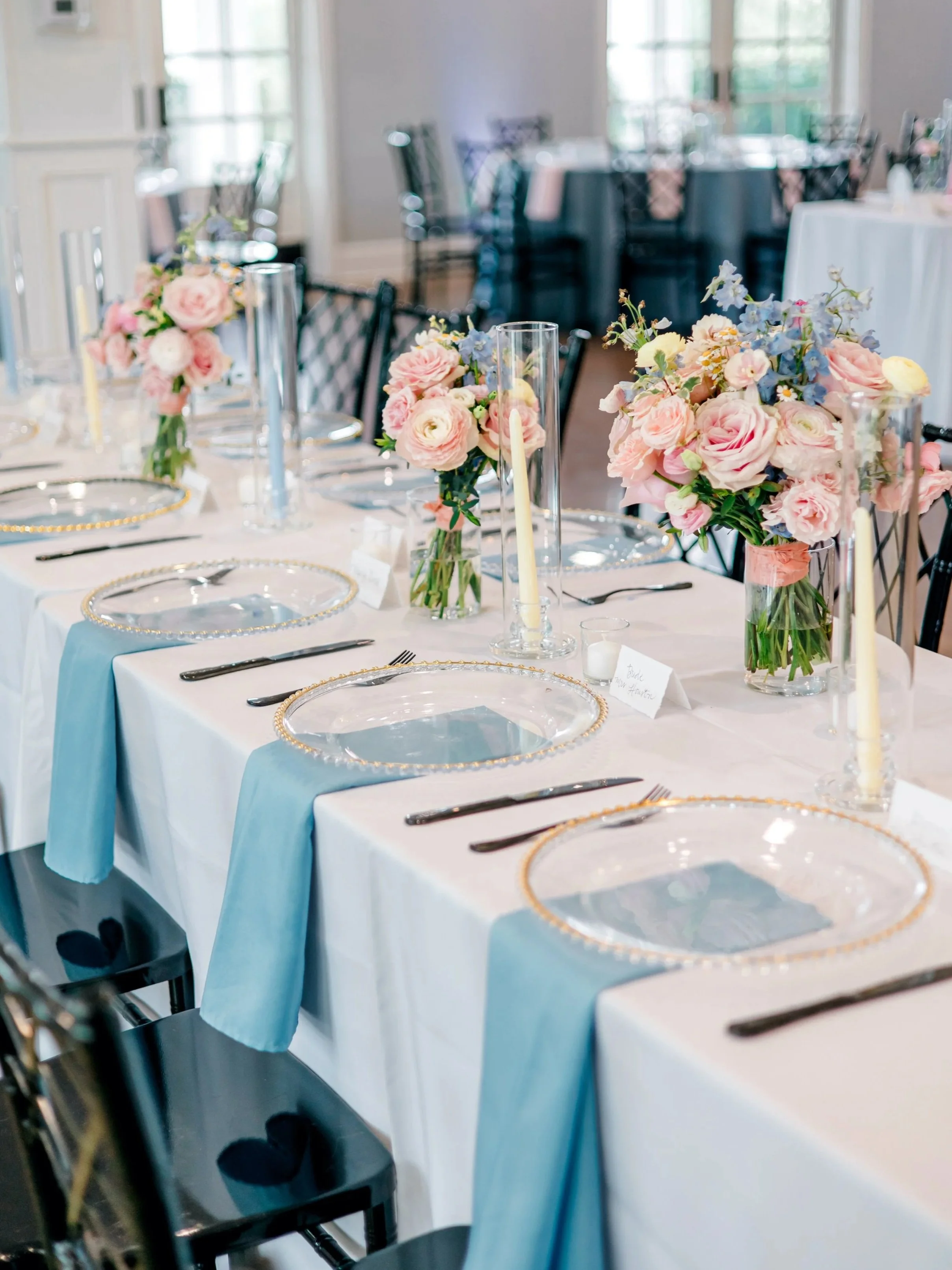 Elegant dining table set with pink and blue floral centerpieces, clear plates with gold beaded rims, black cutlery, tall candles in glass holders, white tablecloths with blue runners in a well-lit room.