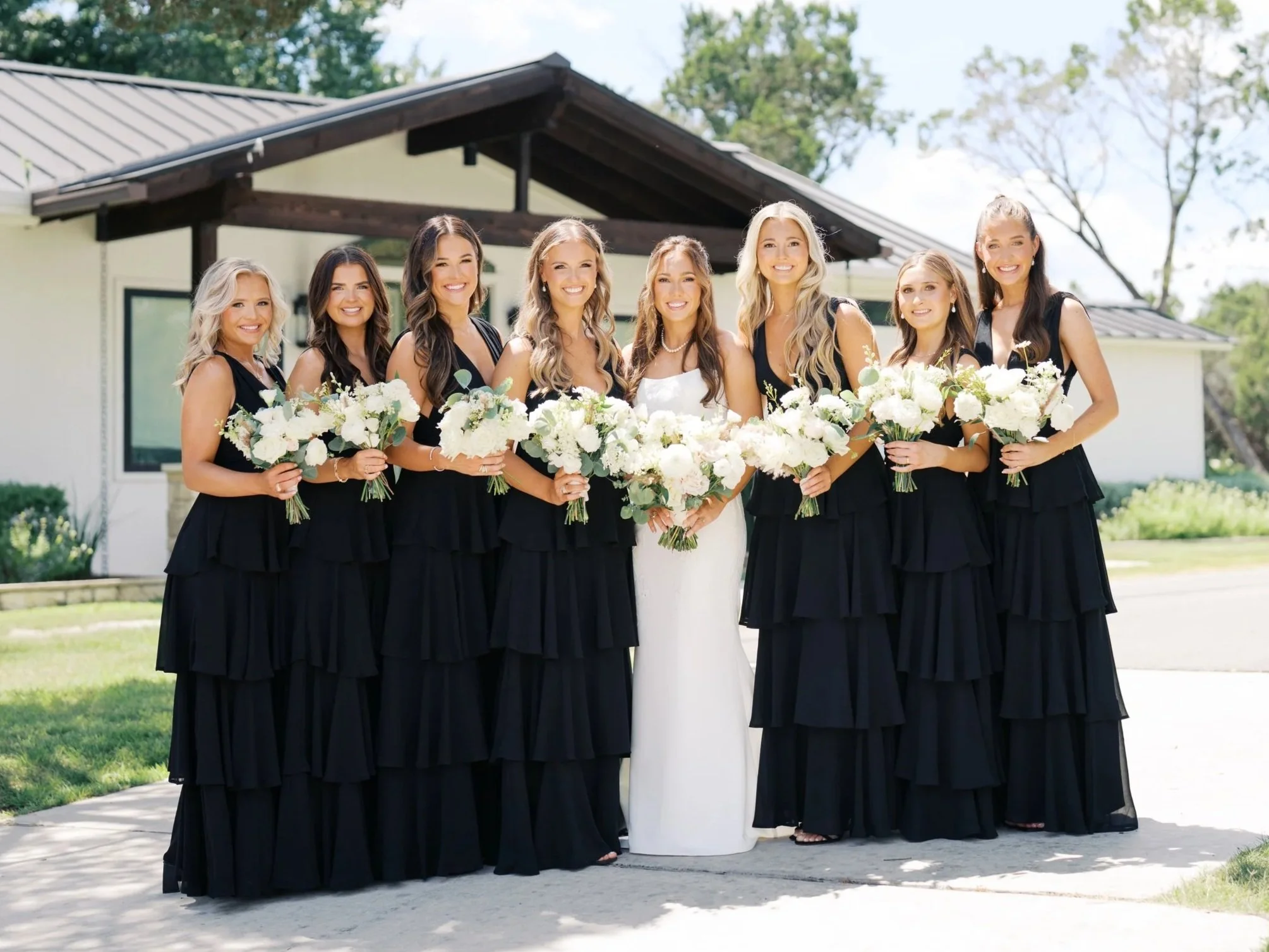 Bride with six bridesmaids outdoors holding white floral bouquets, in front of modern house with trees.