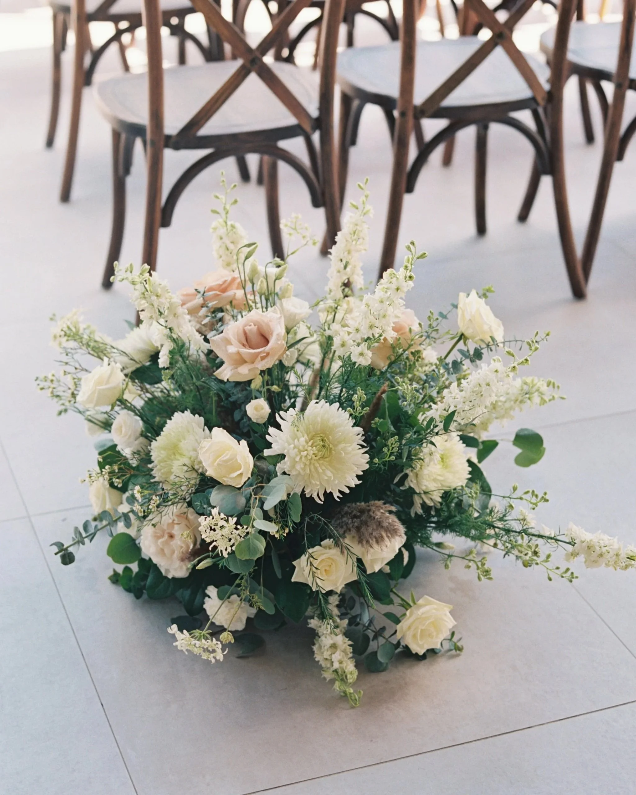 A floral arrangement with white and light pink flowers, including roses and dahlias, placed on the floor with chairs in the background.