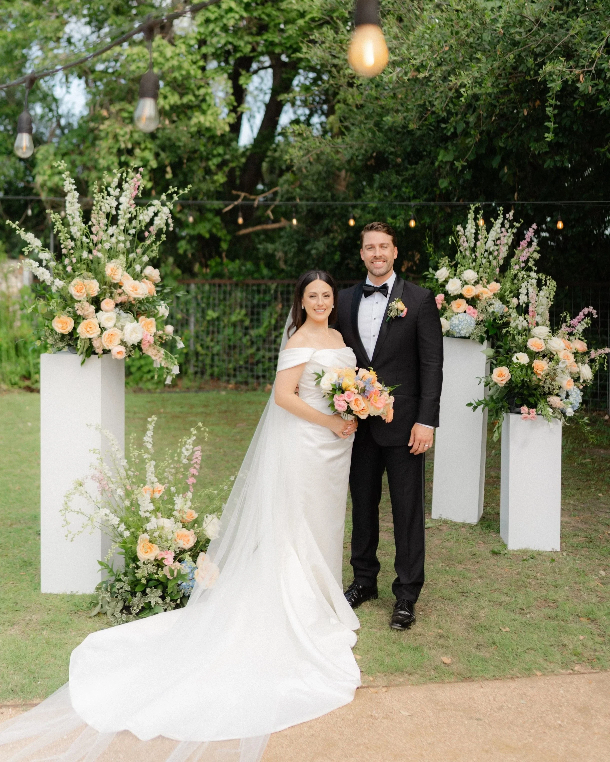 A bride and groom standing outdoors among floral arrangements, smiling, with string lights overhead and trees in the background.