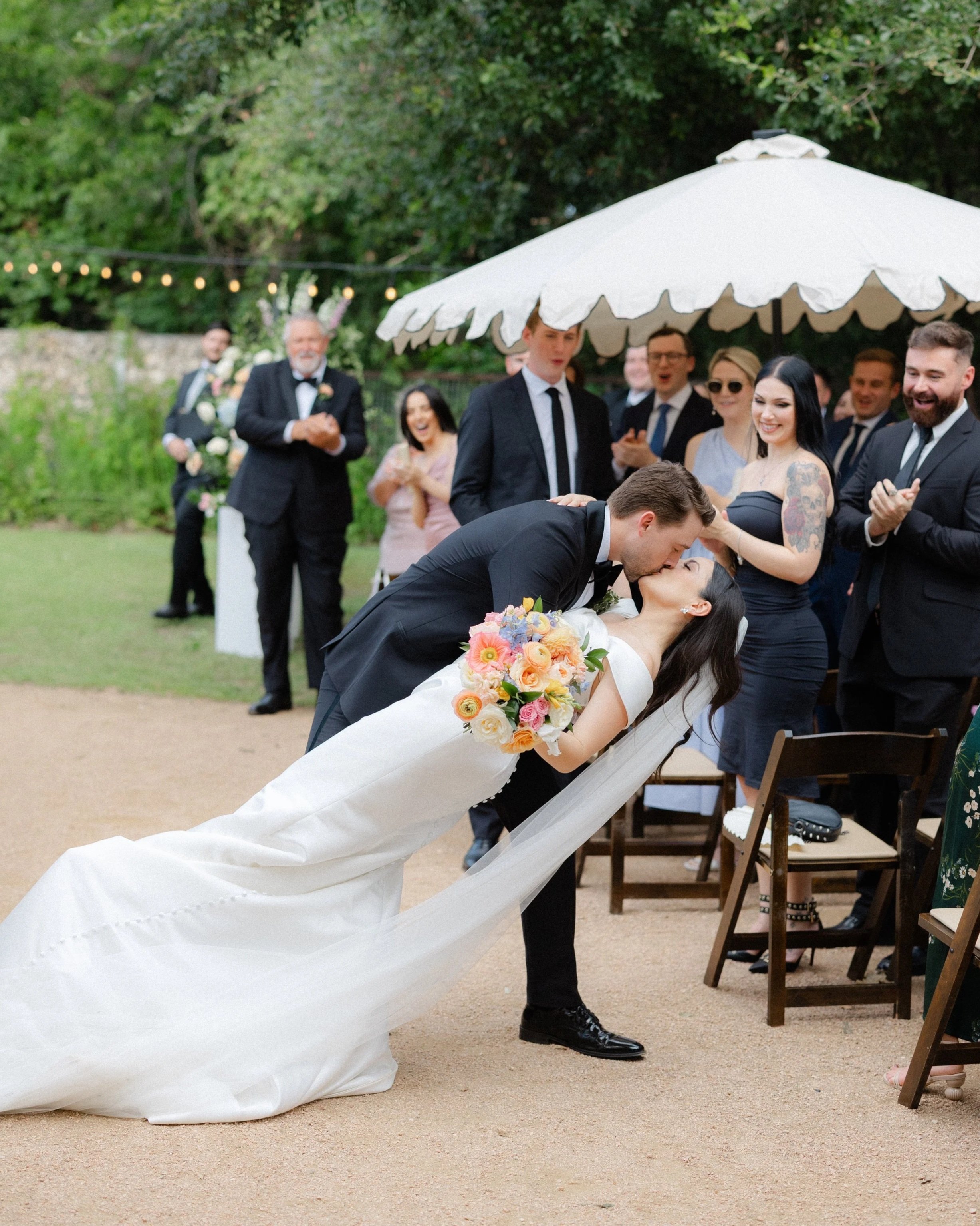 Wedding couple sharing a kiss under a parasol with wedding guests clapping and smiling around them outdoors.