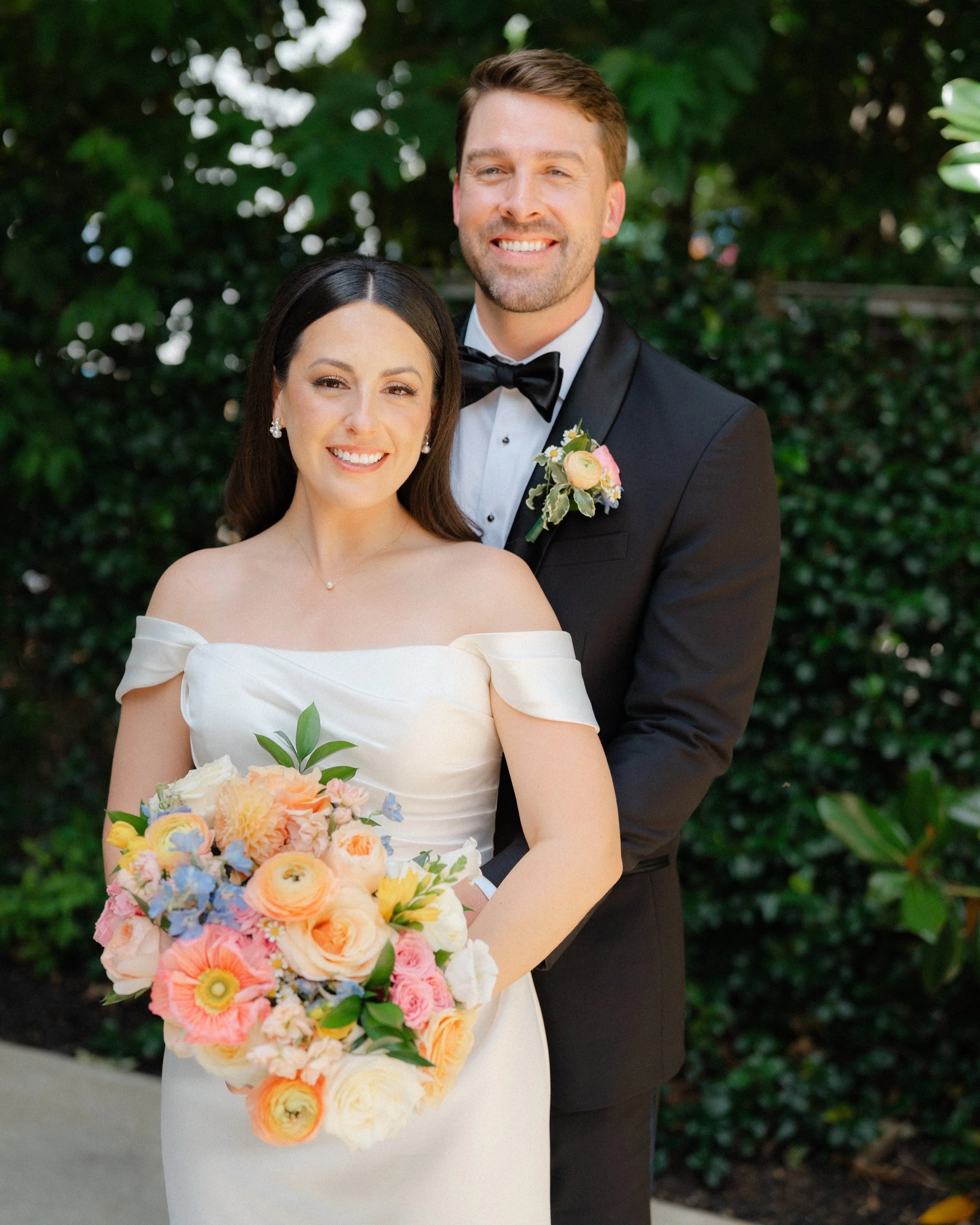 A newly married couple at their wedding, standing outdoors. The bride wears an off-the-shoulder white gown and holding a large colorful bouquet. The groom is dressed in a black tuxedo with a bow tie and a flower boutonniere on his lapel. They are smi