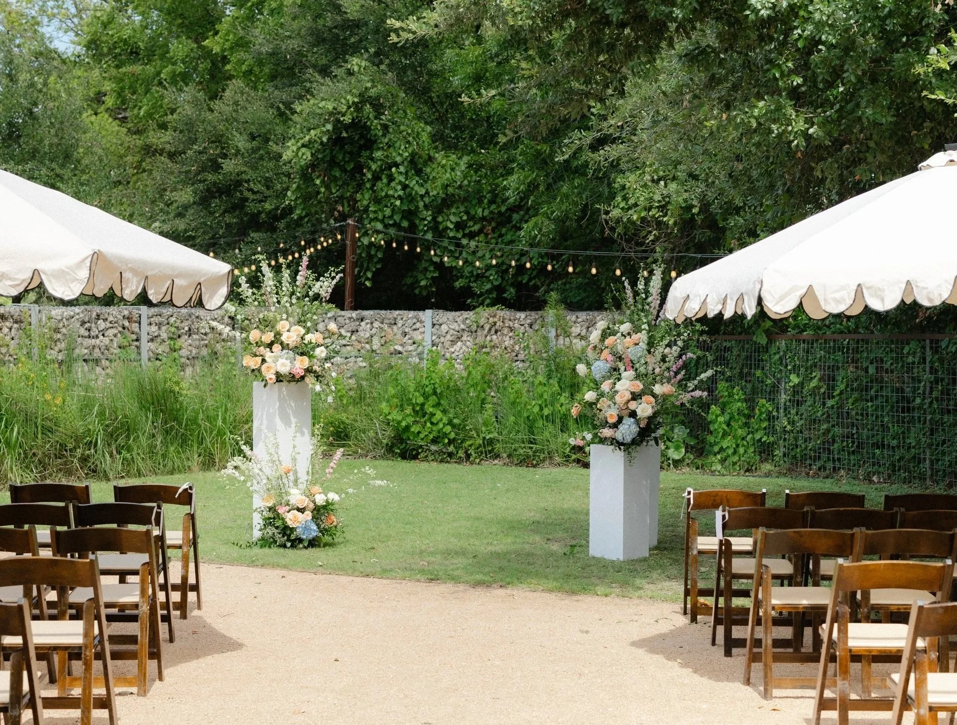 Outdoor wedding ceremony setup with two floral arrangements on white pedestals, seats arranged on both sides, white tent canopies, greenery in background, and string lights overhead.