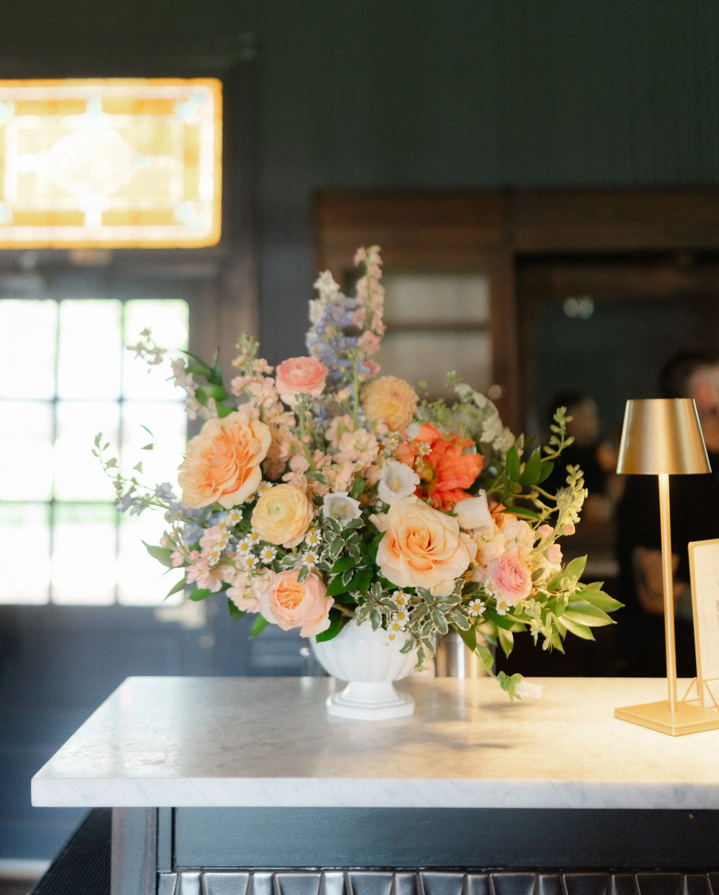 A large bouquet of colorful flowers in a white vase on a table, with a gold desk lamp nearby, in a well-lit interior setting.