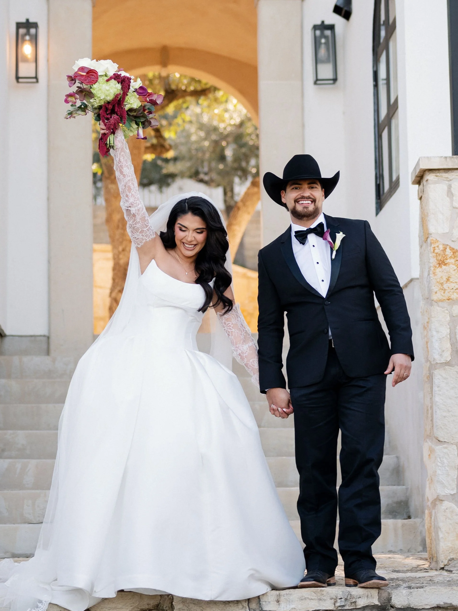 A bride and groom holding hands and smiling after their wedding, standing on stone steps outside a building with arched entryway and lantern-style lights.