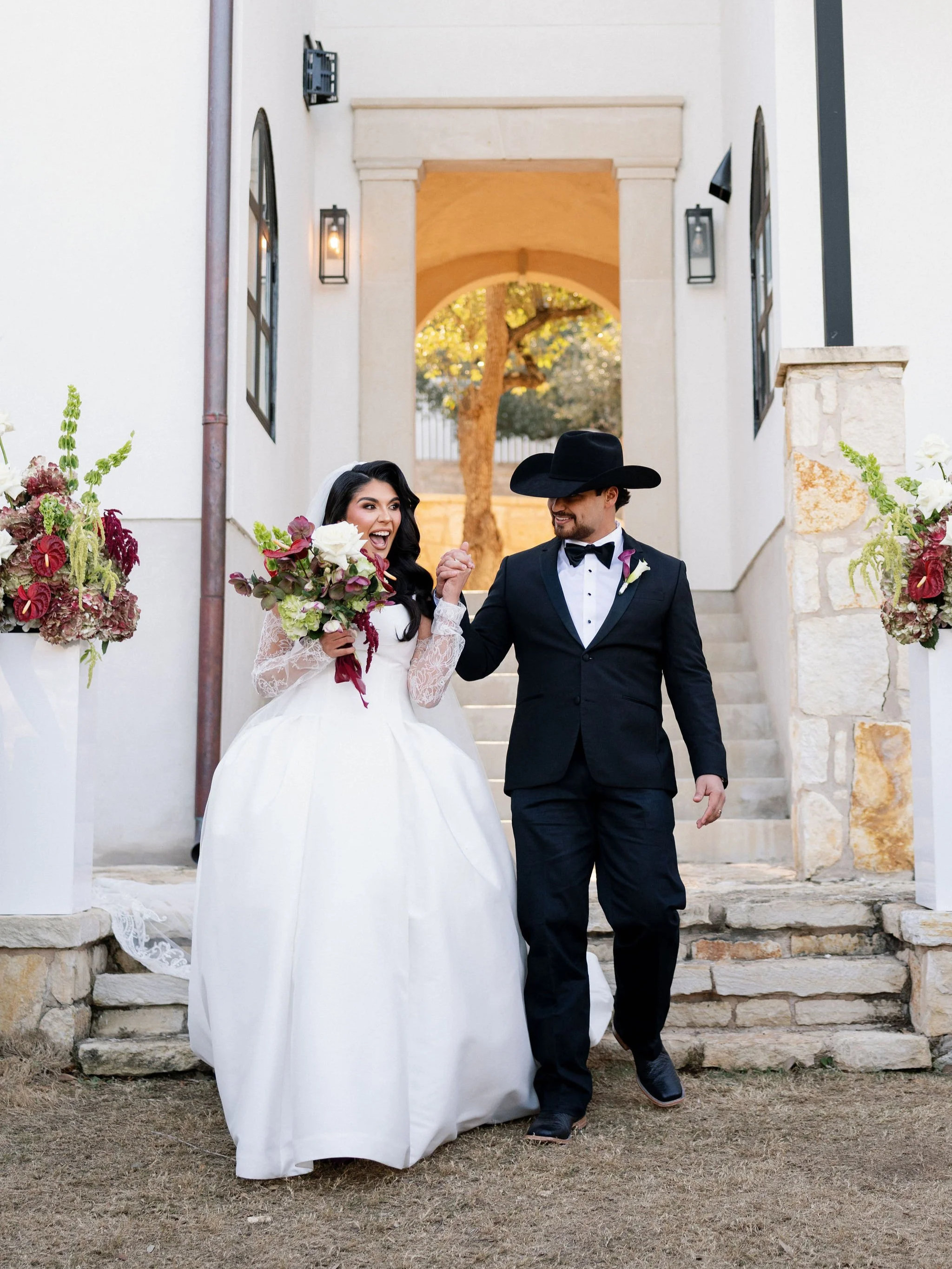 A bride in a white wedding dress holding a bouquet, smiling excitedly, walking arm-in-arm with a groom in a black suit and cowboy hat, on stairs outside a venue decorated with floral arrangements.
