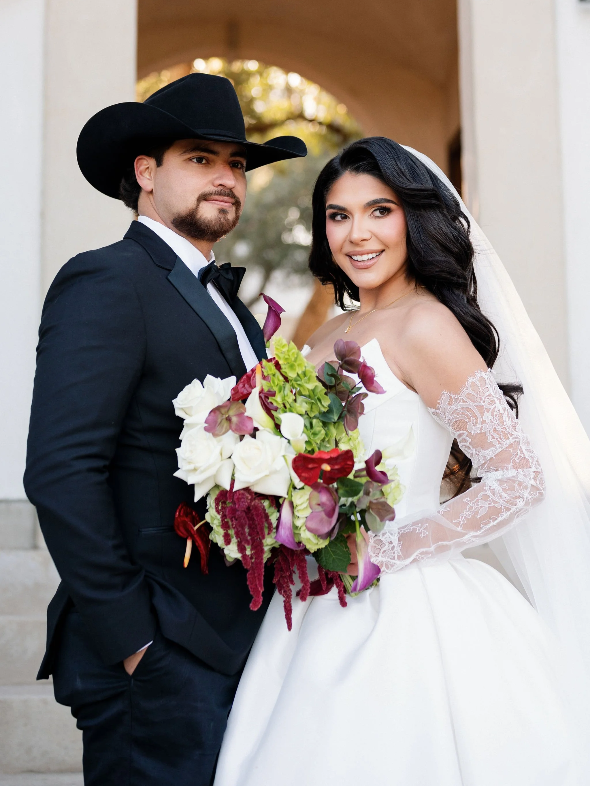 A bride and groom in wedding attire standing together outdoors, the bride holding a colorful bouquet, both smiling at the camera.