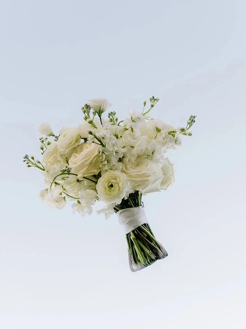 A white bridal bouquet with roses and other white flowers, wrapped with a white ribbon.