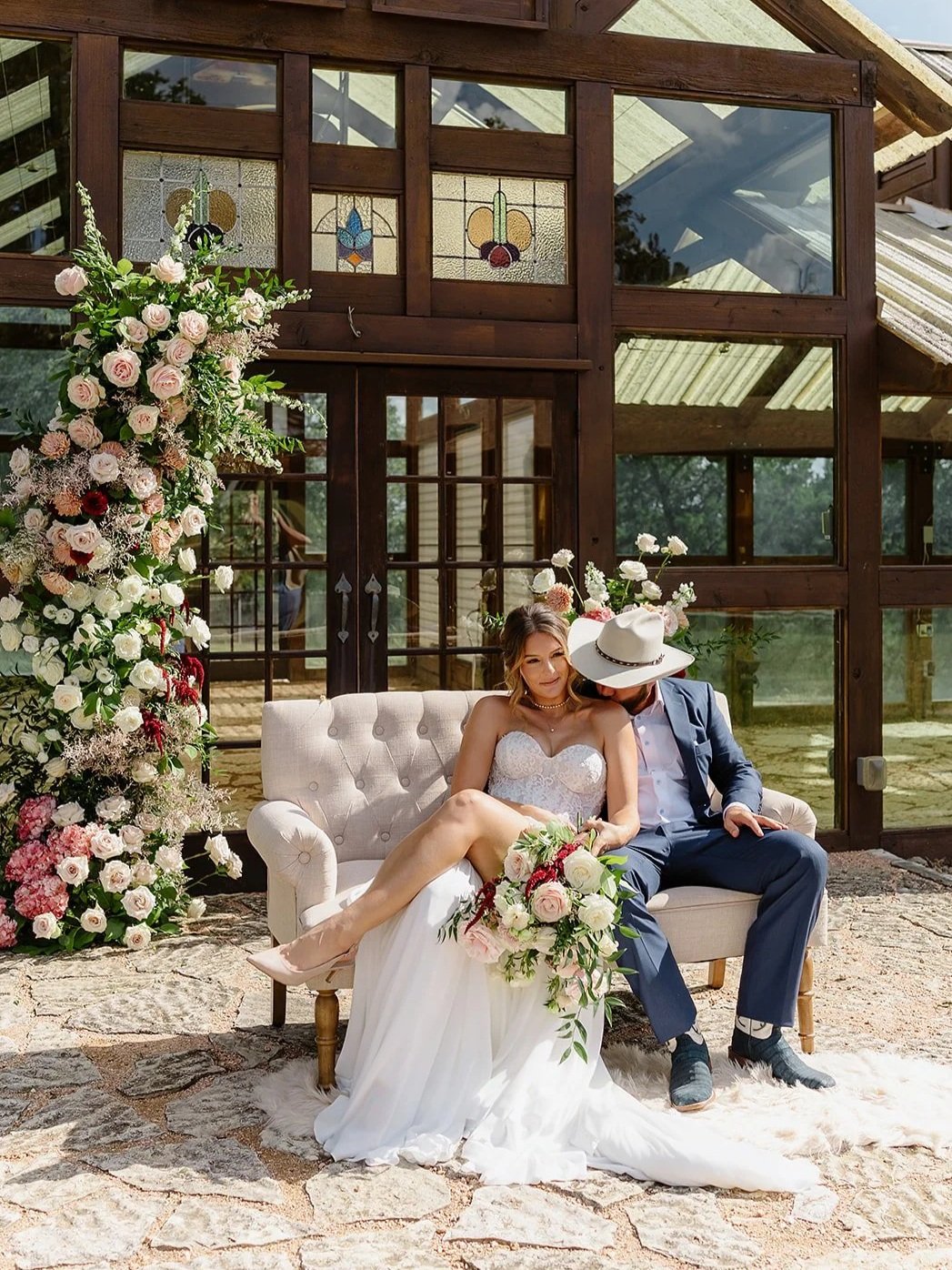 A bride and groom sitting on a cream-colored loveseat outdoors, surrounded by a large floral arrangement, in front of a wooden building with glass windows and decorative stained glass panels.