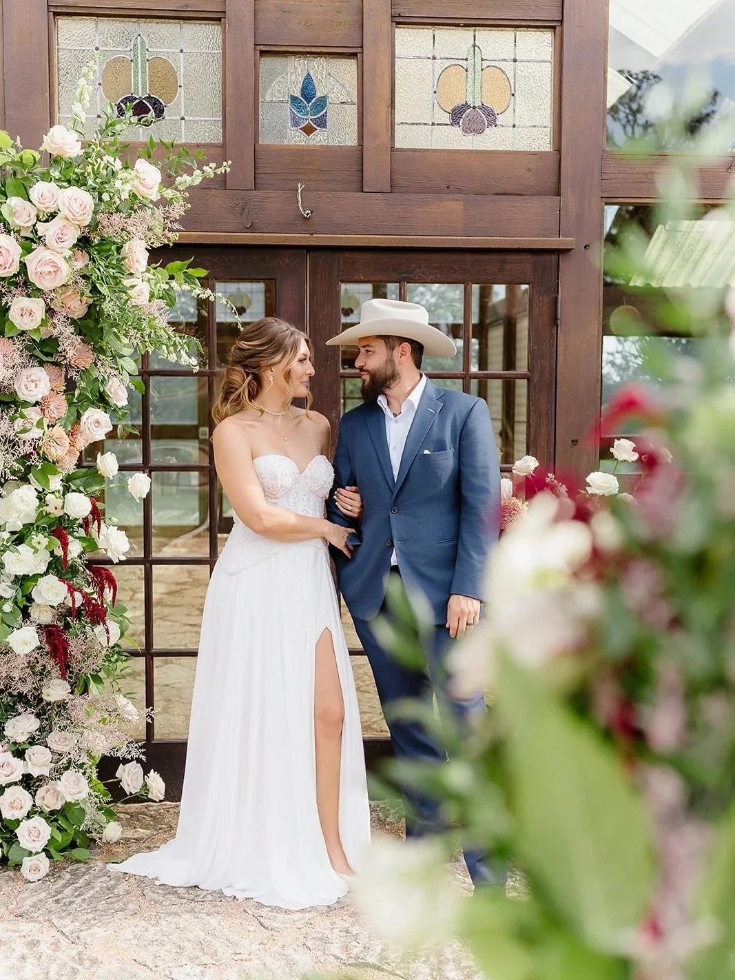 A bride and groom standing close together, smiling at each other during their wedding ceremony, with a floral arch and stained glass window behind them.