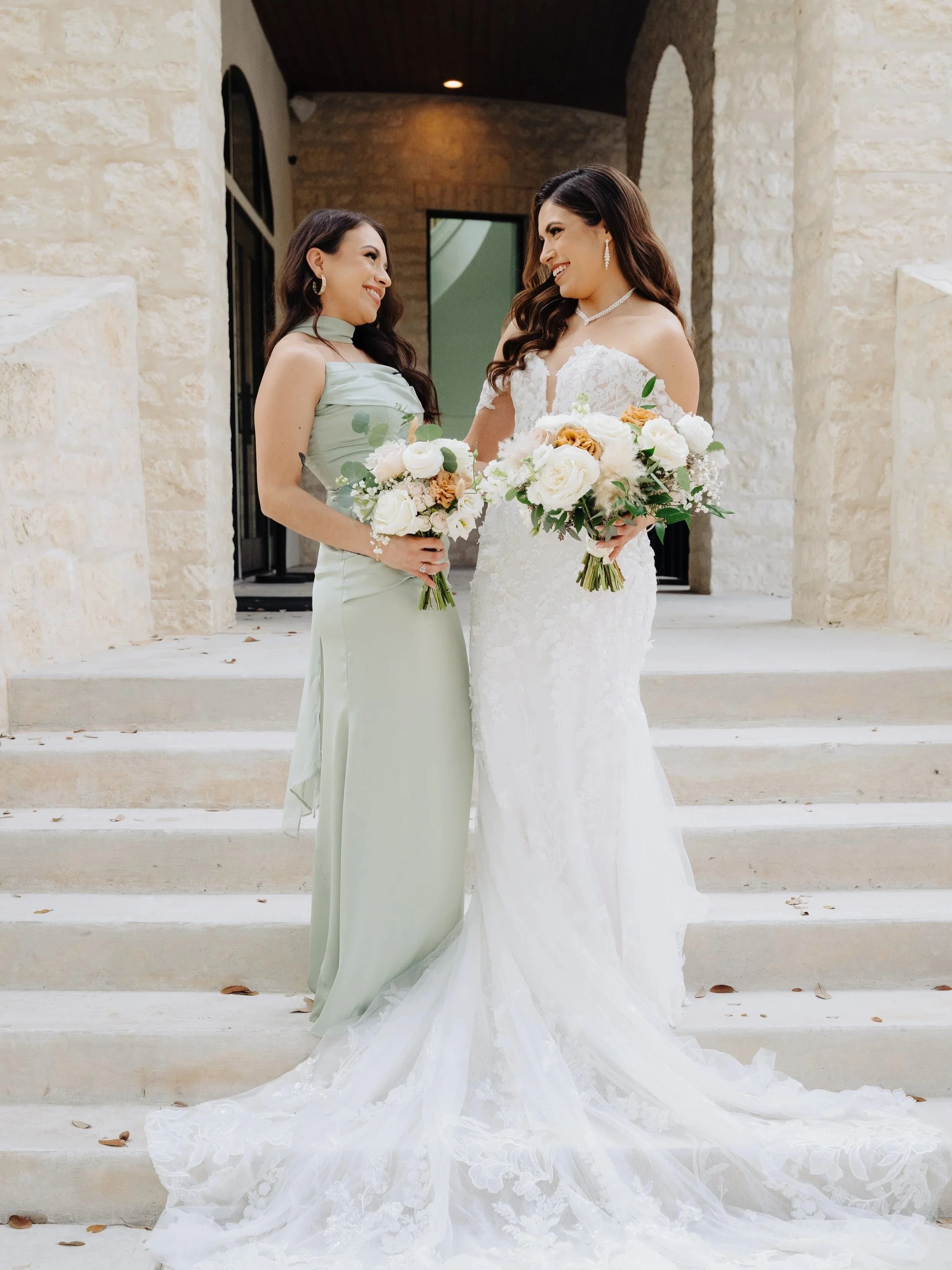 A bride in a white lace wedding dress holding a bouquet of white and peach flowers standing on steps with a woman in a light green dress holding a similar bouquet, smiling at each other outside a stone building.