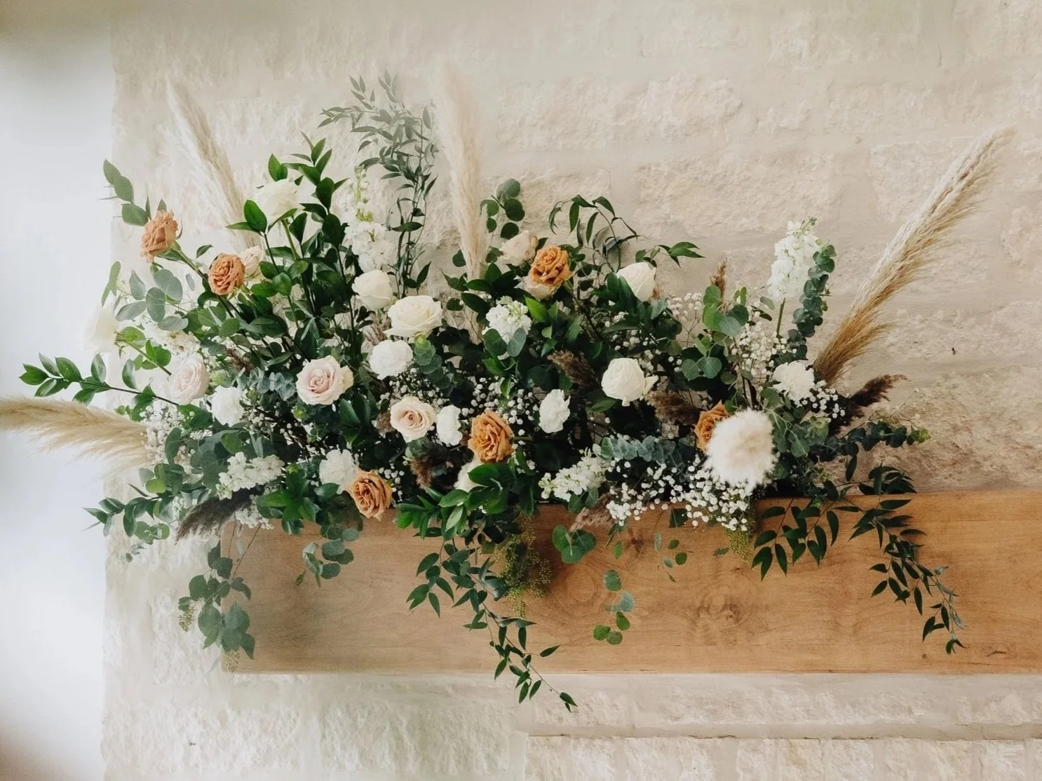 A floral arrangement in a rectangular wooden container with white, peach, and brown flowers, greenery, and dried pampas grass, placed against a textured beige wall.