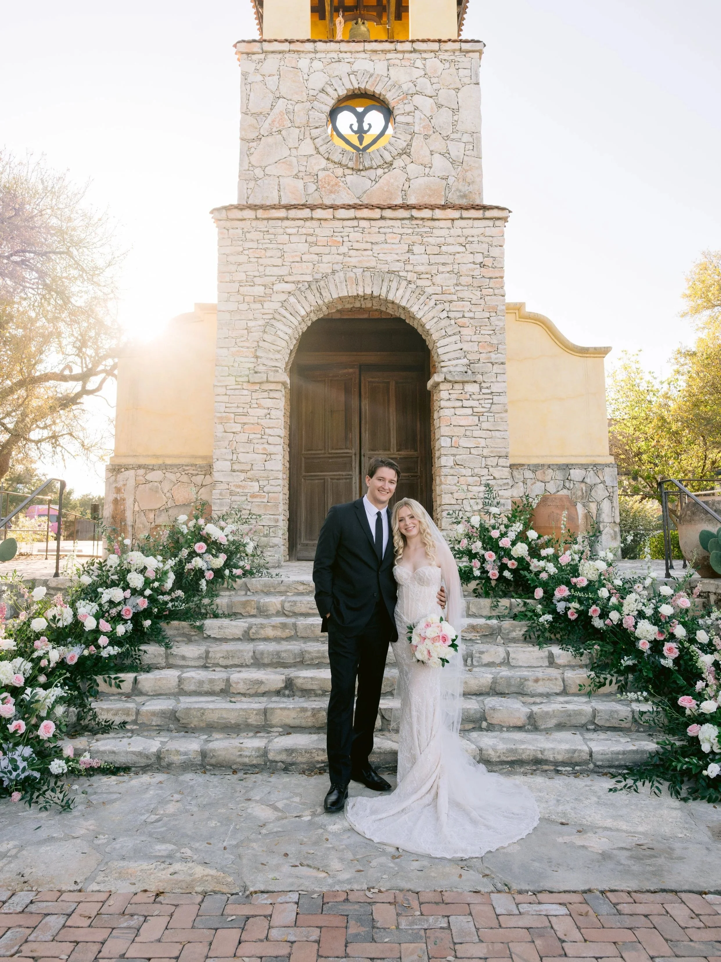 A newlywed couple stands on stone steps in front of a rustic stone church, surrounded by floral arrangements, with the bride holding a bouquet, and both smiling happily.
