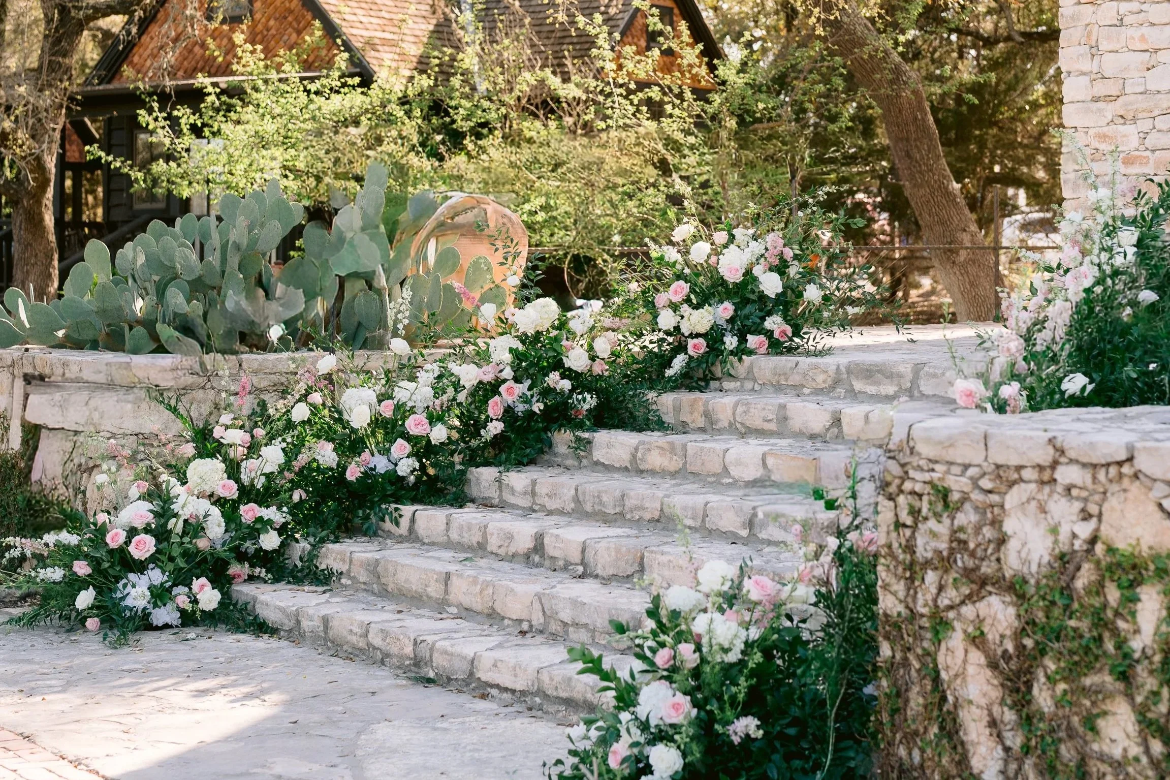 Stone staircase decorated with pink and white flowers, surrounded by succulents and greenery, leading to a rustic house with a brick roof.