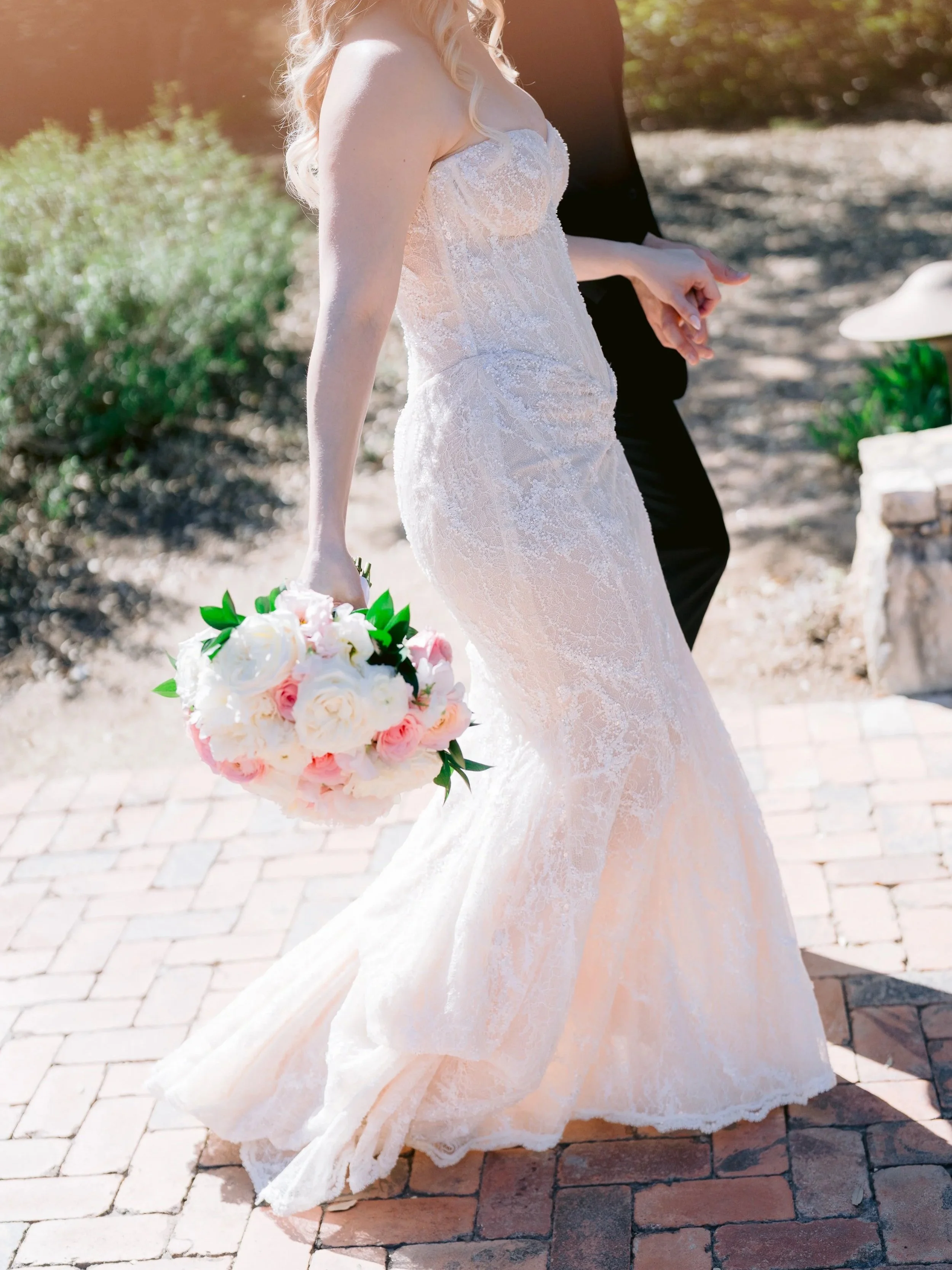 A bride in a strapless lace wedding gown holding a bouquet of pink and white roses, standing outdoors on a brick path.