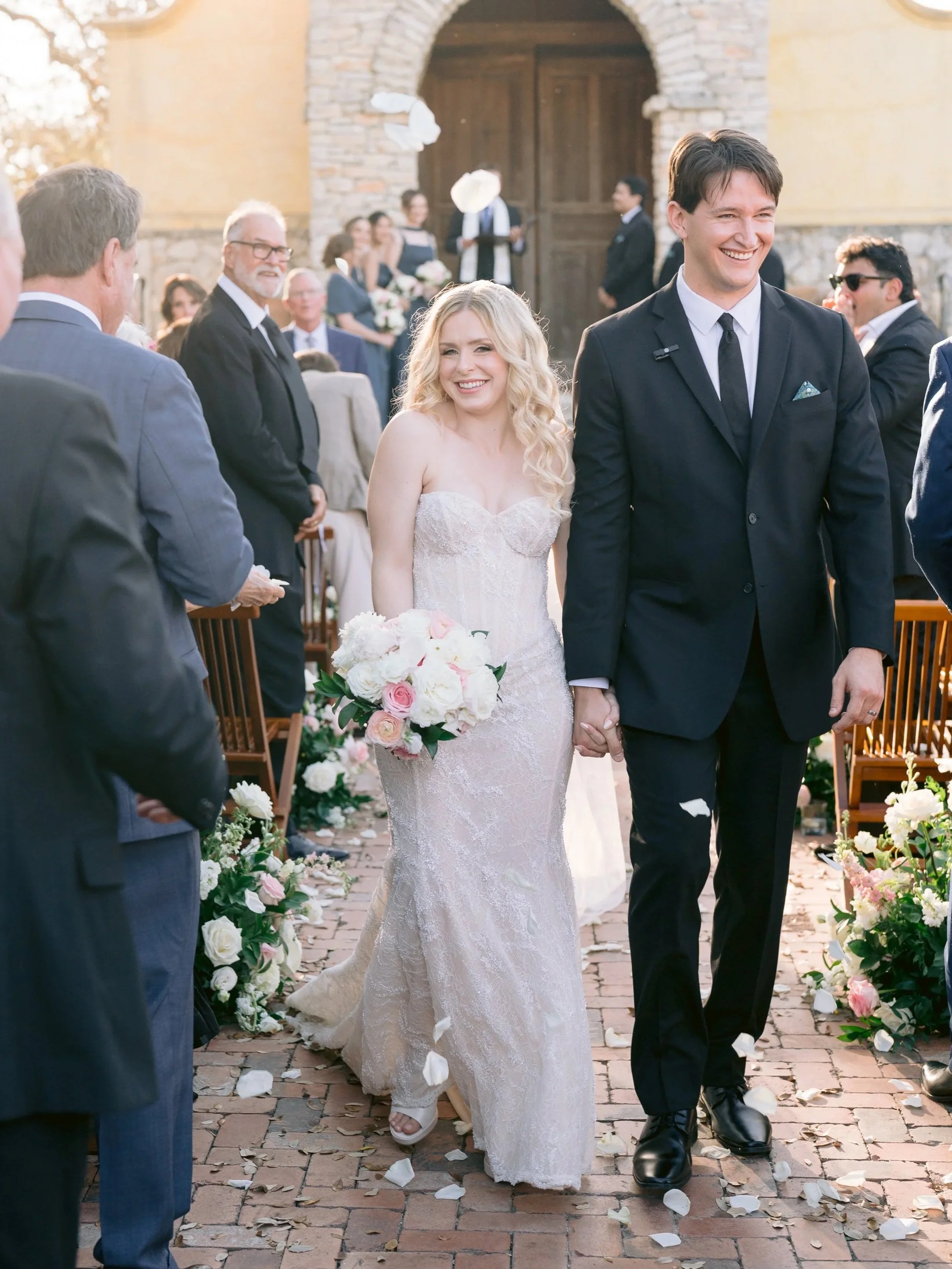 A newlywed couple walking hand-in-hand down an outdoor aisle, surrounded by friends and family, with the bride in a lace wedding dress holding a bouquet of white and pink roses, and the groom in a black suit and tie, during a wedding celebration.