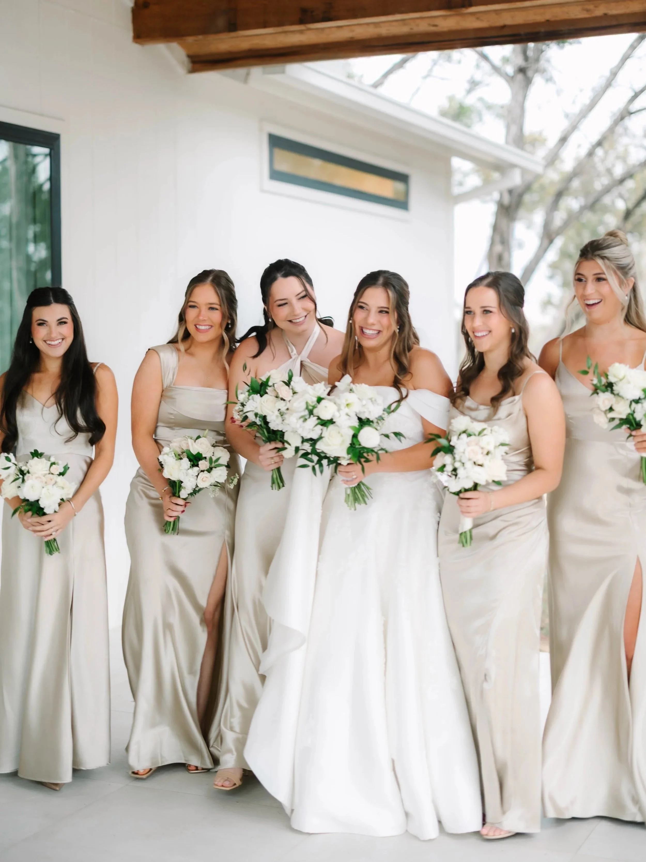 A group of six women in bridesmaid dresses and wedding dress holding bouquets, standing together and smiling in front of a modern house exterior.