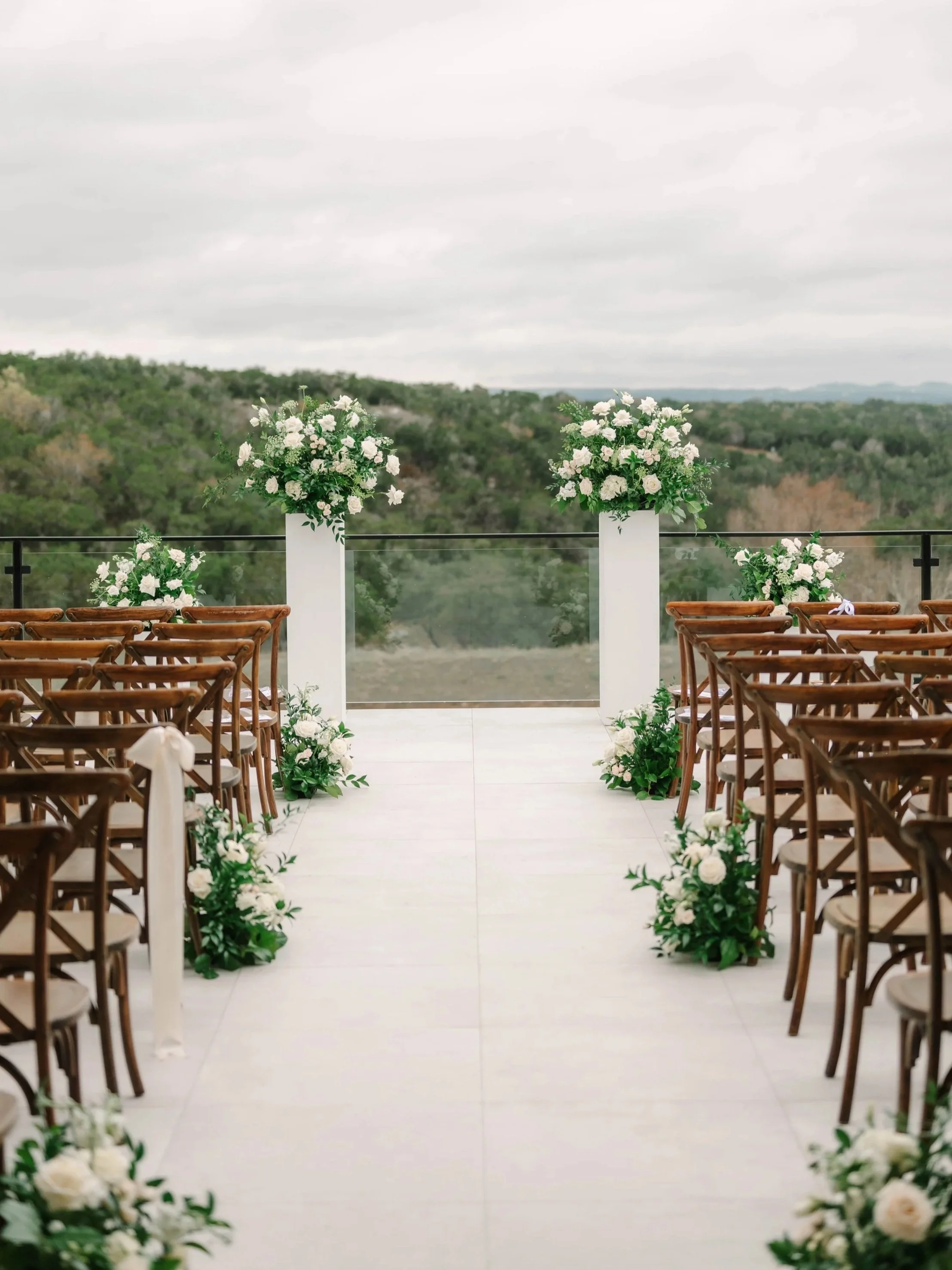 An outdoor wedding ceremony setup with wooden chairs and white floral arrangements, overlooking a scenic view of hills and trees under a cloudy sky.