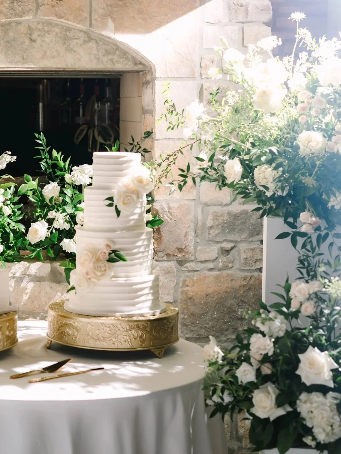 A wedding cake with white ridges decorated with white roses and green leaves, placed on a gold stand, surrounded by white floral arrangements on a table.