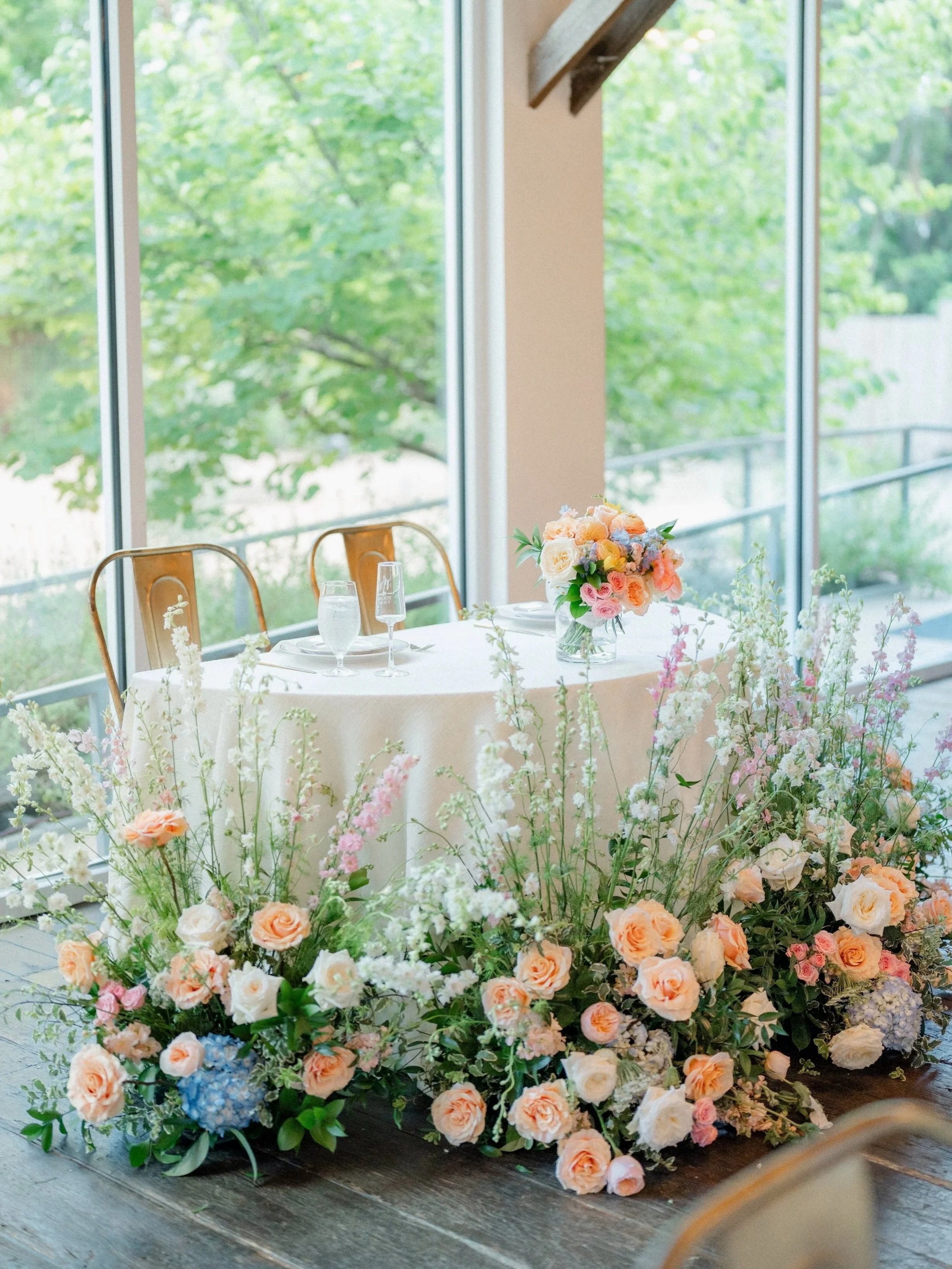 Wedding reception table with floral centerpiece and cascading flowers in front, set near large windows with green trees outside.