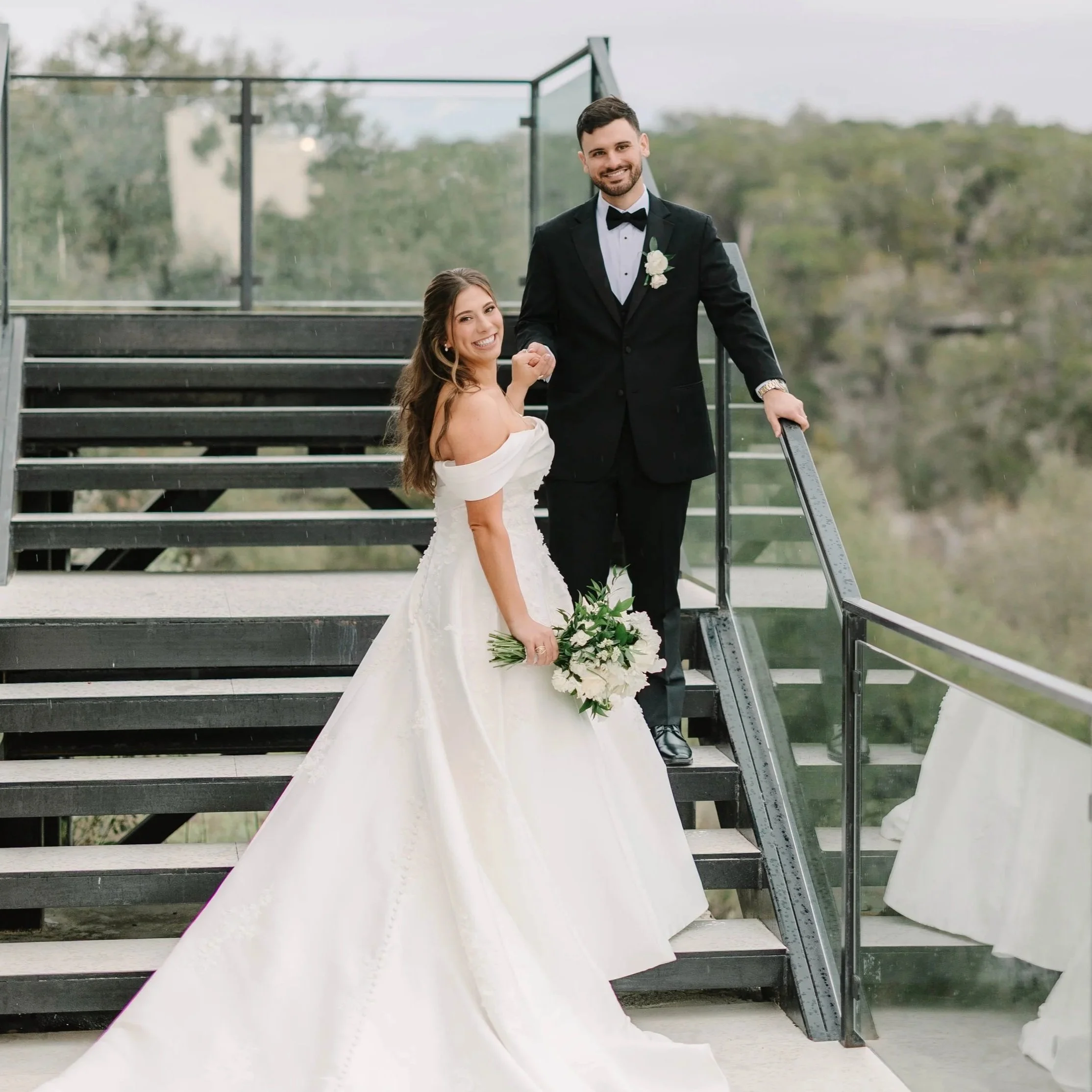 A bride and groom stand on a glass staircase outdoors, smiling at the camera; the bride holds a bouquet of white flowers, and the groom is in a black tuxedo with a bow tie and boutonniere.