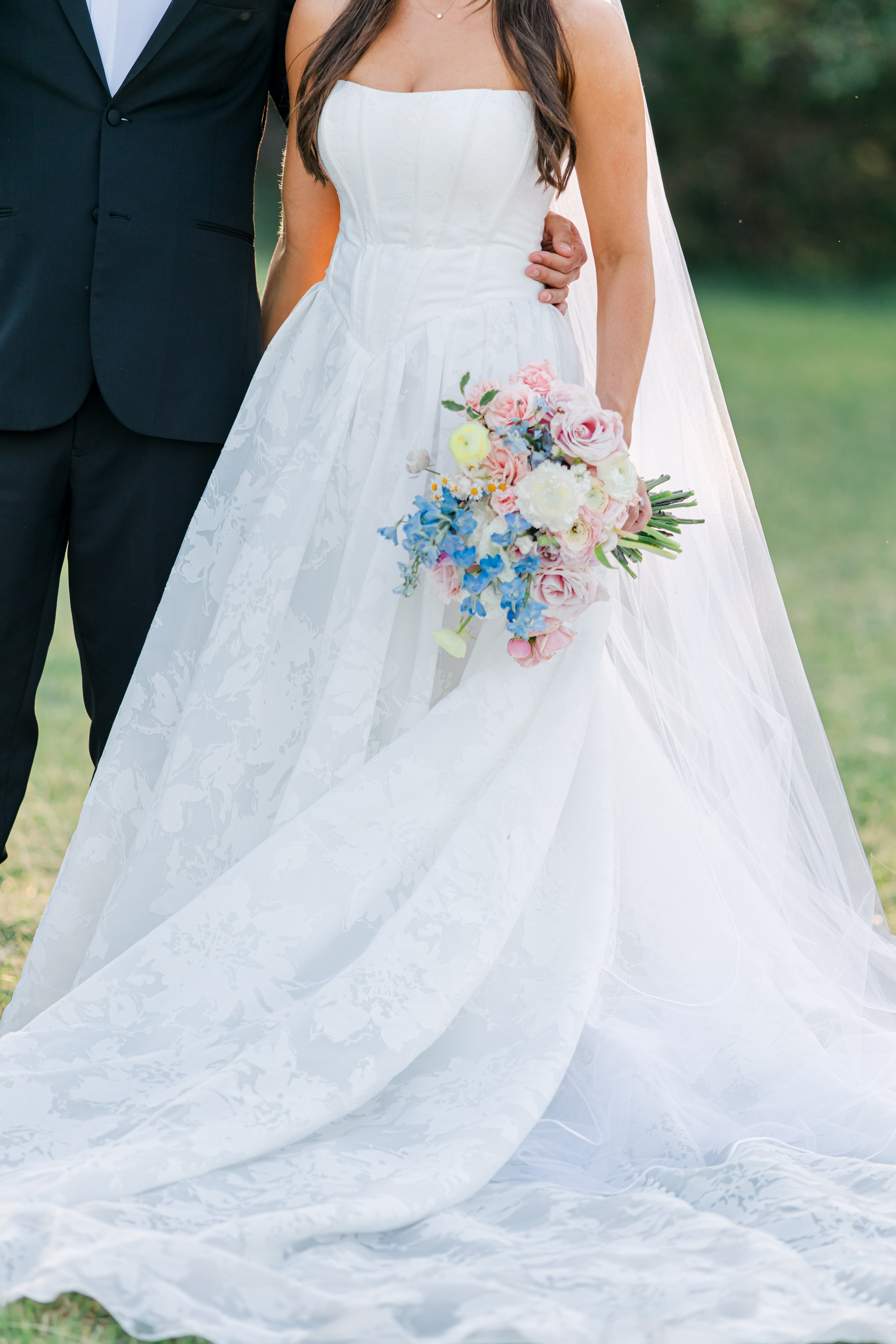 Bride in a white wedding gown holding a bouquet of pink, white, yellow, and blue flowers, standing outdoors with a groom partly visible beside her.