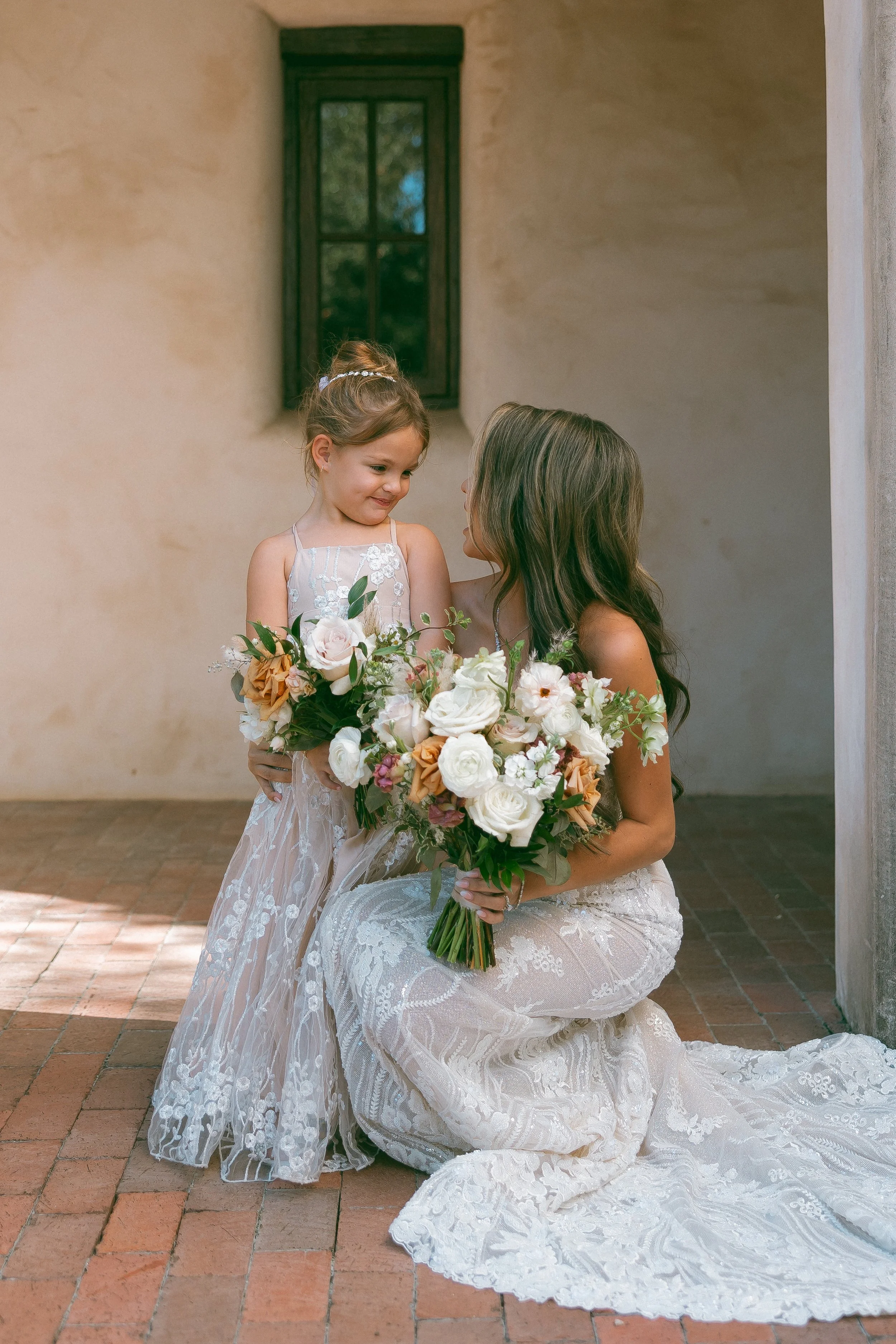 A woman in a lace dress kneeling on the brick floor, holding a bouquet of white, peach, and pink flowers, talking to a young girl in a white dress with floral embroidery, also holding a bouquet of similar flowers, in front of a rustic wall with a win