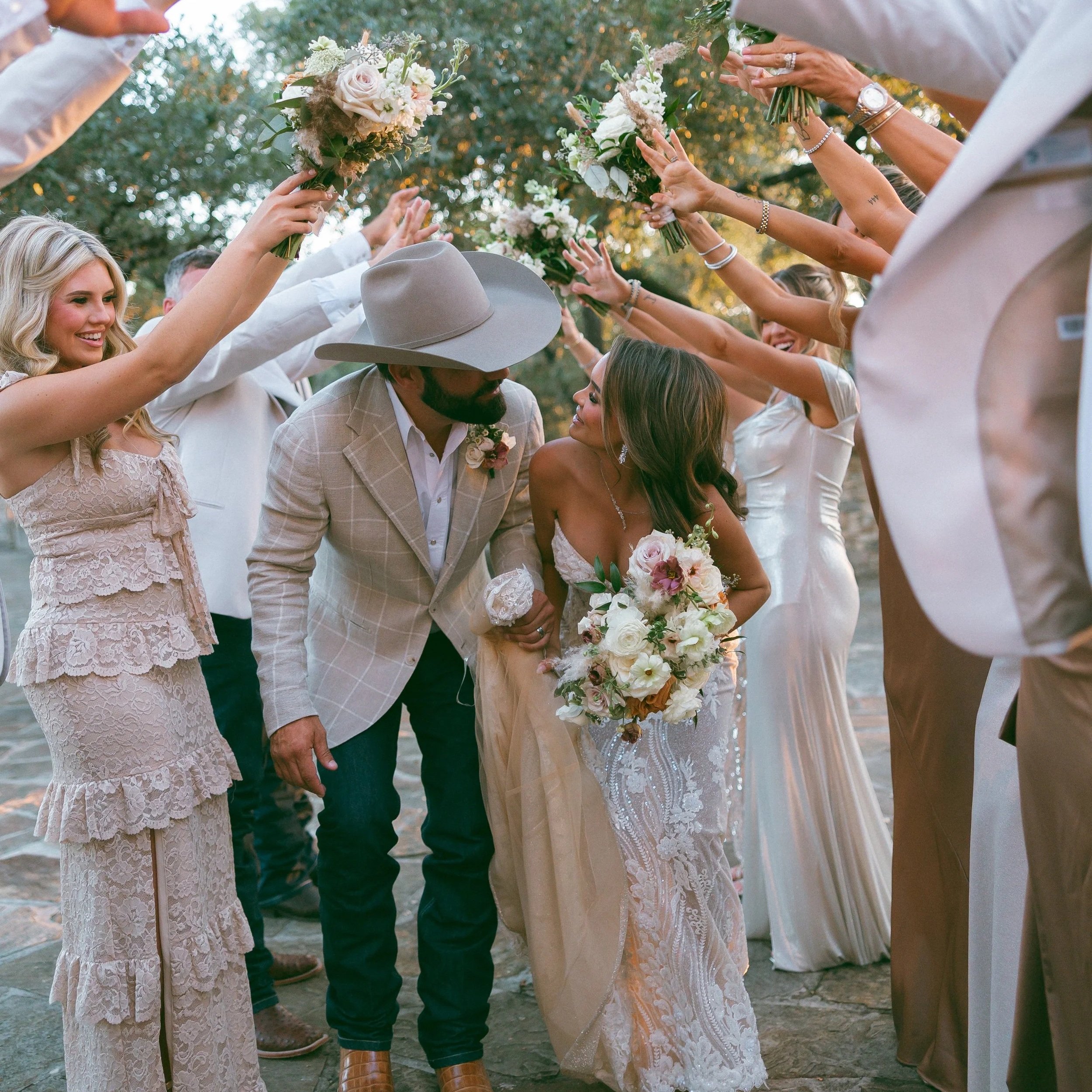 Bride and groom surrounded by friends holding bouquets, celebrating outdoors.