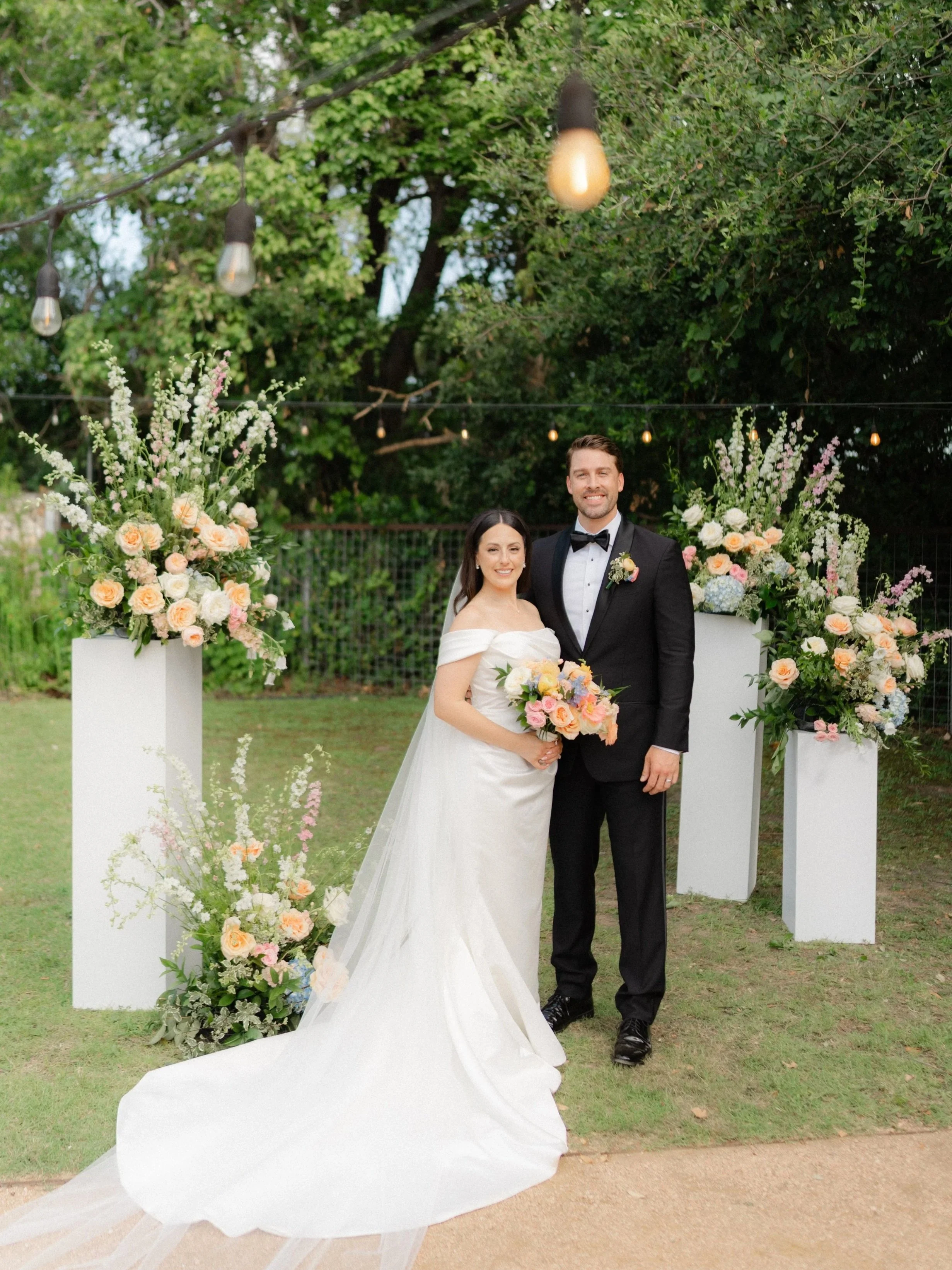 A bride and groom standing together outdoors in front of a floral arch, smiling at the camera, with string lights overhead and greenery in the background.