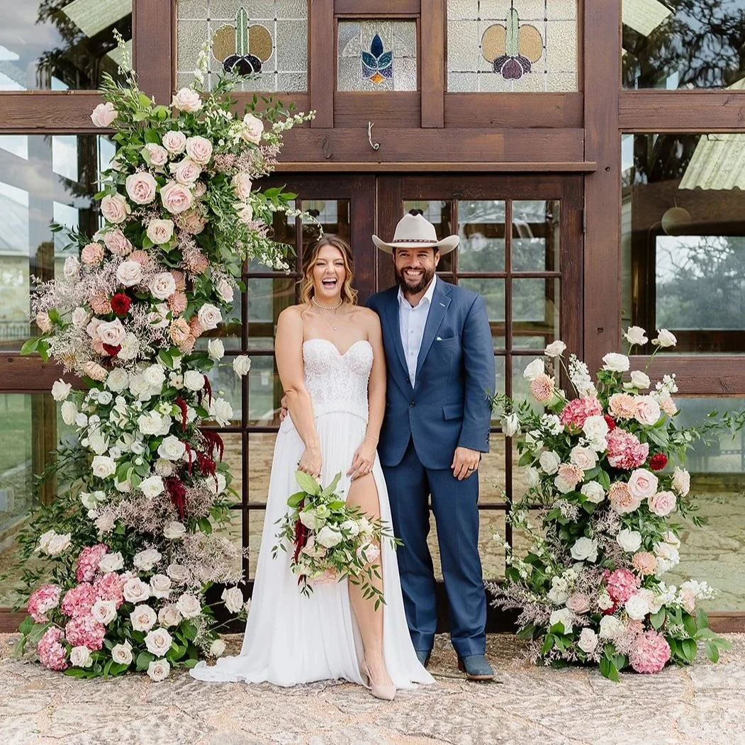 A smiling bride in a white wedding dress holding a bouquet, standing next to a groom in a blue suit and cowboy hat, in front of a wooden backdrop decorated with pink and white flowers.