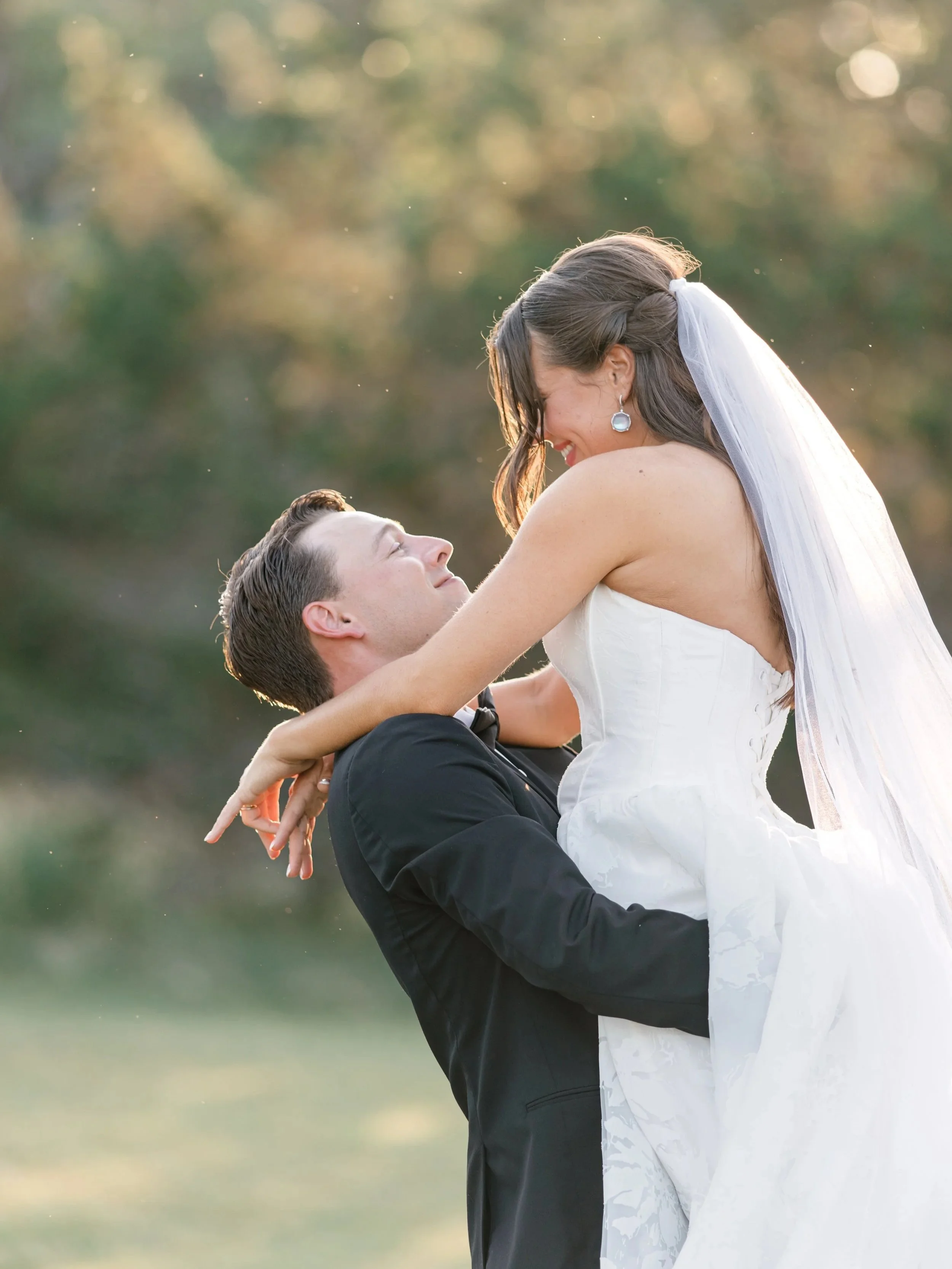 Bride and groom embracing outdoors during sunset, with the groom lifting the bride who is smiling.