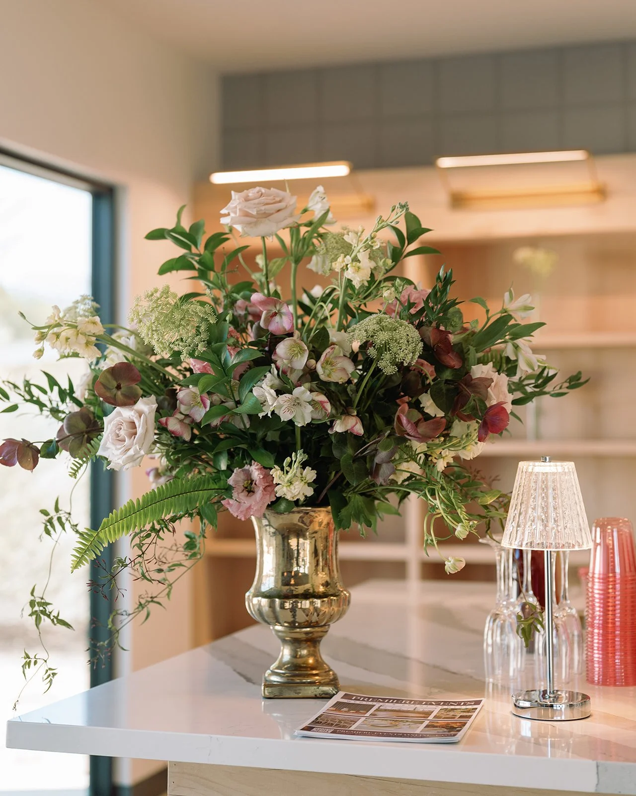 A large bouquet of assorted pink, white, and green flowers in a shiny metallic vase on a white table with a small lamp, glassware, pink cups, and a magazine.