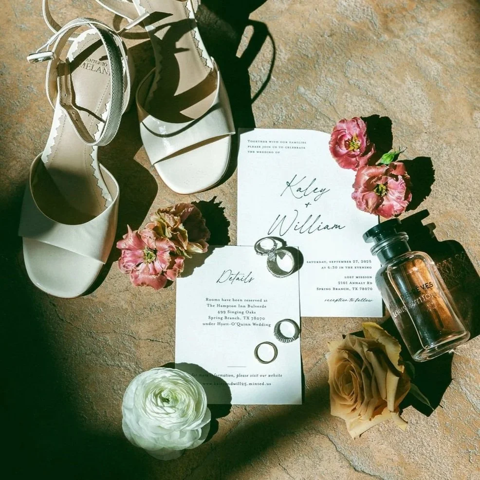 Wedding invitation, white high-heeled shoes, pink and beige roses, wedding rings, perfume bottle, and scattered flower petals on a textured surface.