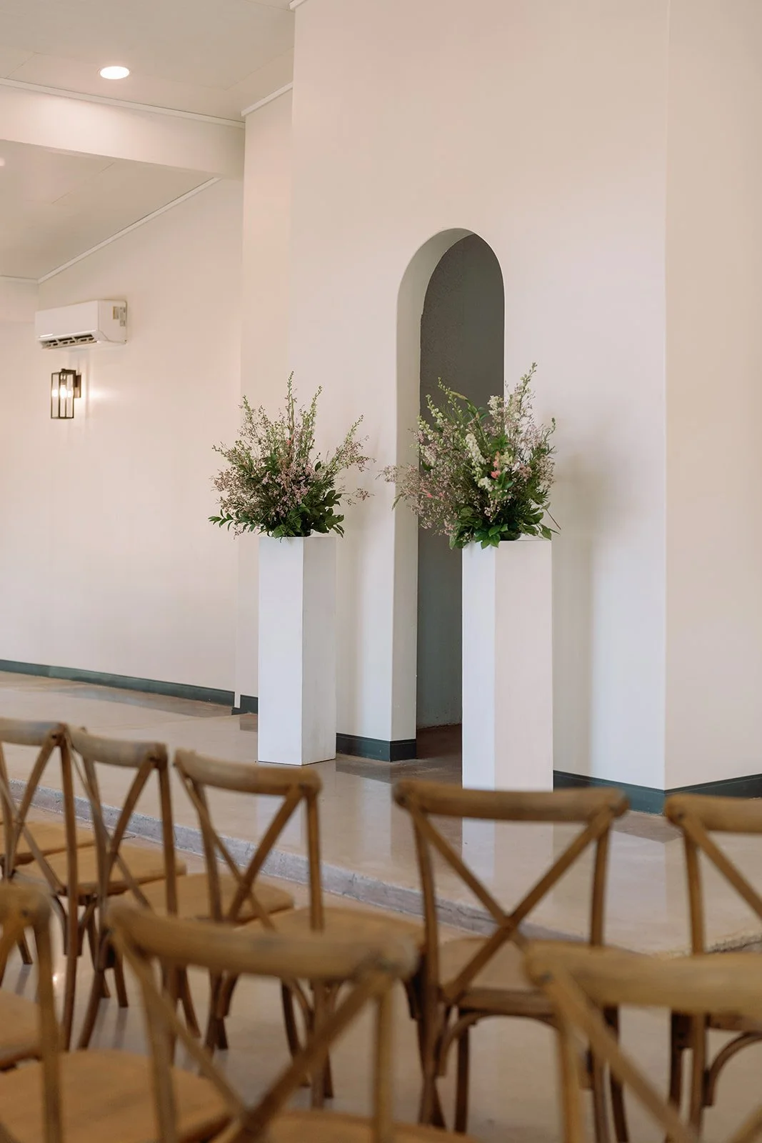 Two white pedestals with floral arrangements placed in front of a wall with an arched opening, in a room with wooden chairs arranged in the foreground.