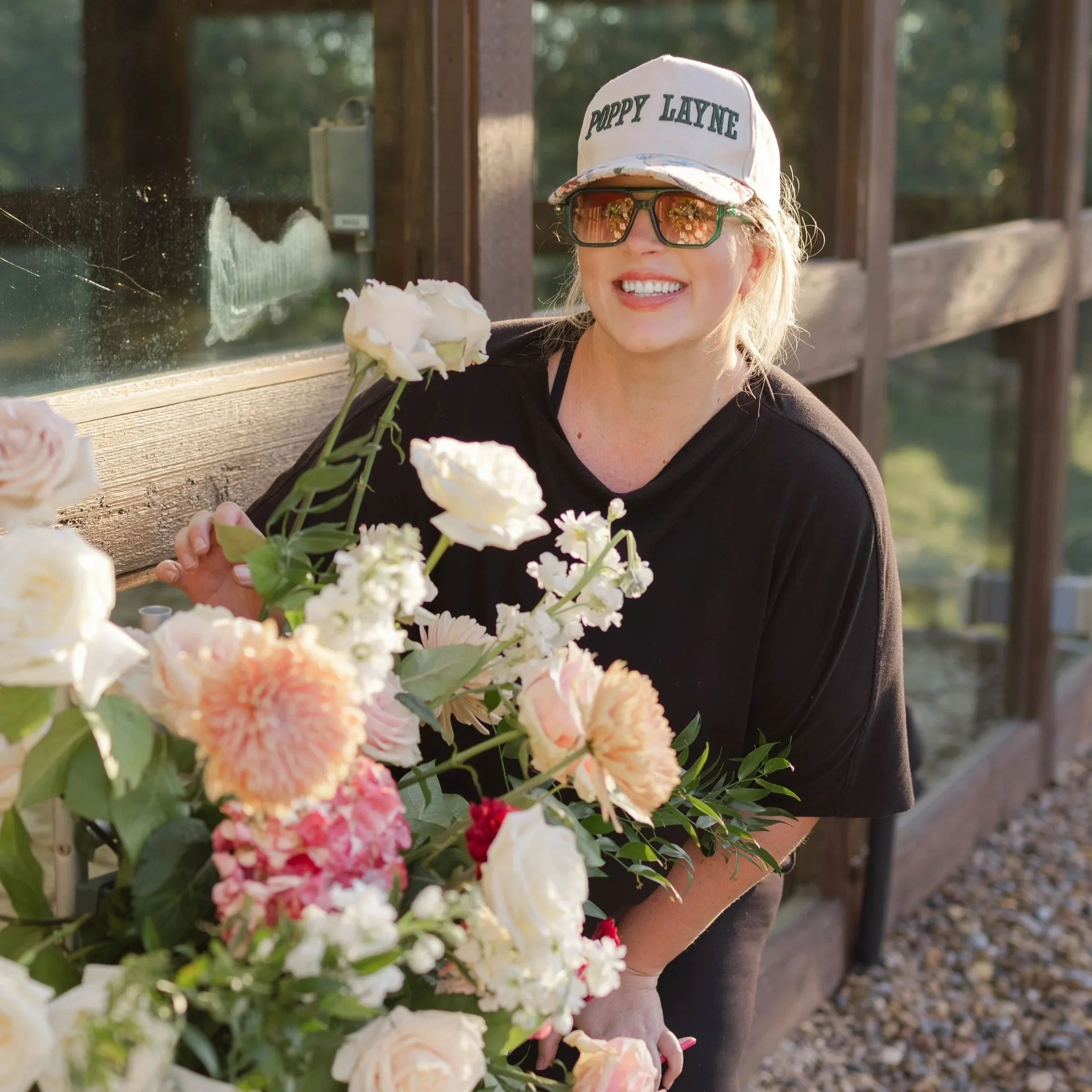 A woman with blonde hair, sunglasses, and a white cap that says 'Poppy Layne' is smiling and posing with a large bouquet of pink and white flowers outdoors near a wooden railing.