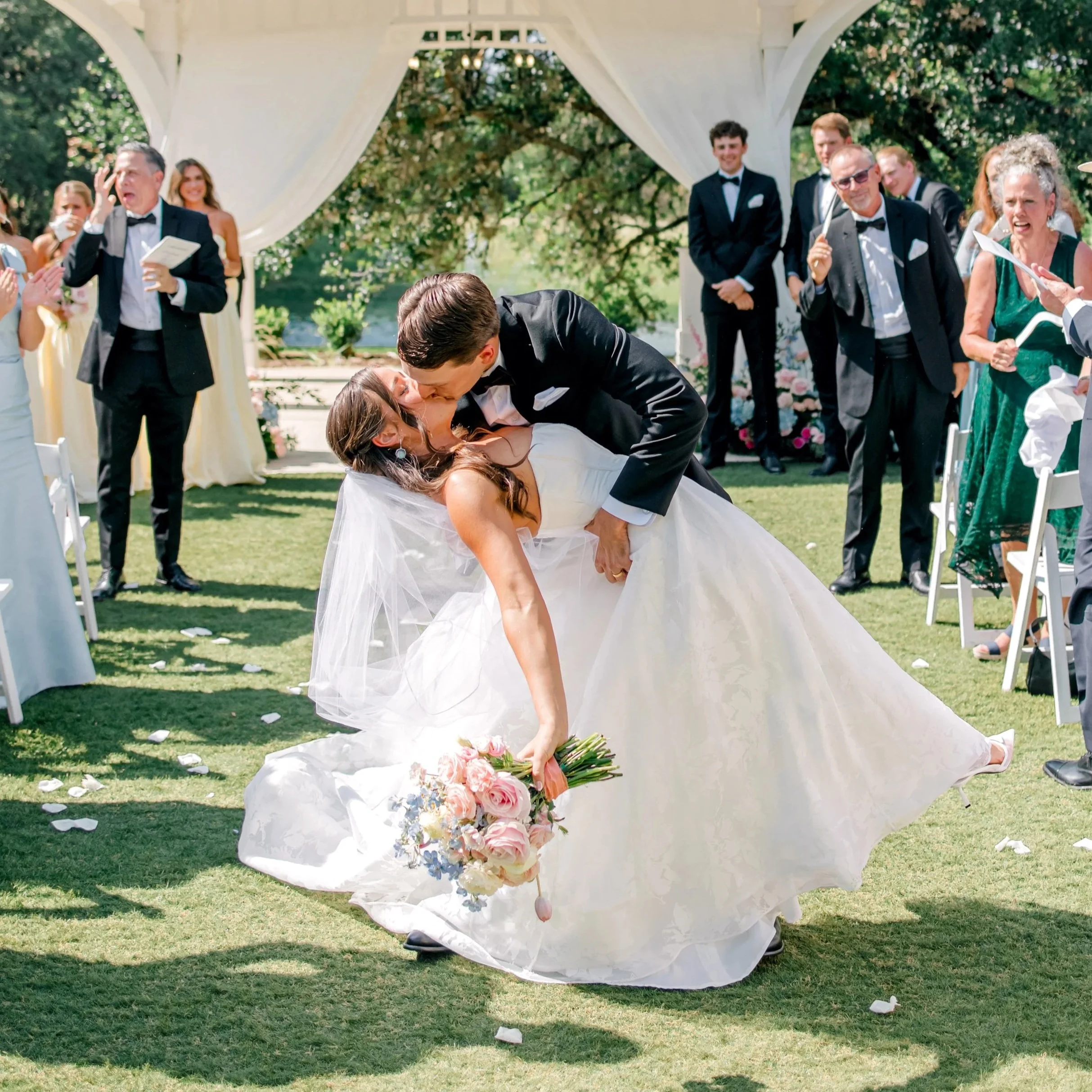 A bride and groom kiss during their wedding ceremony outdoors, with guests watching and celebrating around them.