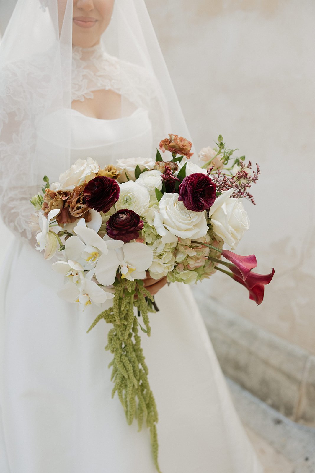 Bridal dress with lace details and veil, holding a bouquet of white, burgundy, pink, and purple flowers including roses, orchids, and calla lilies.