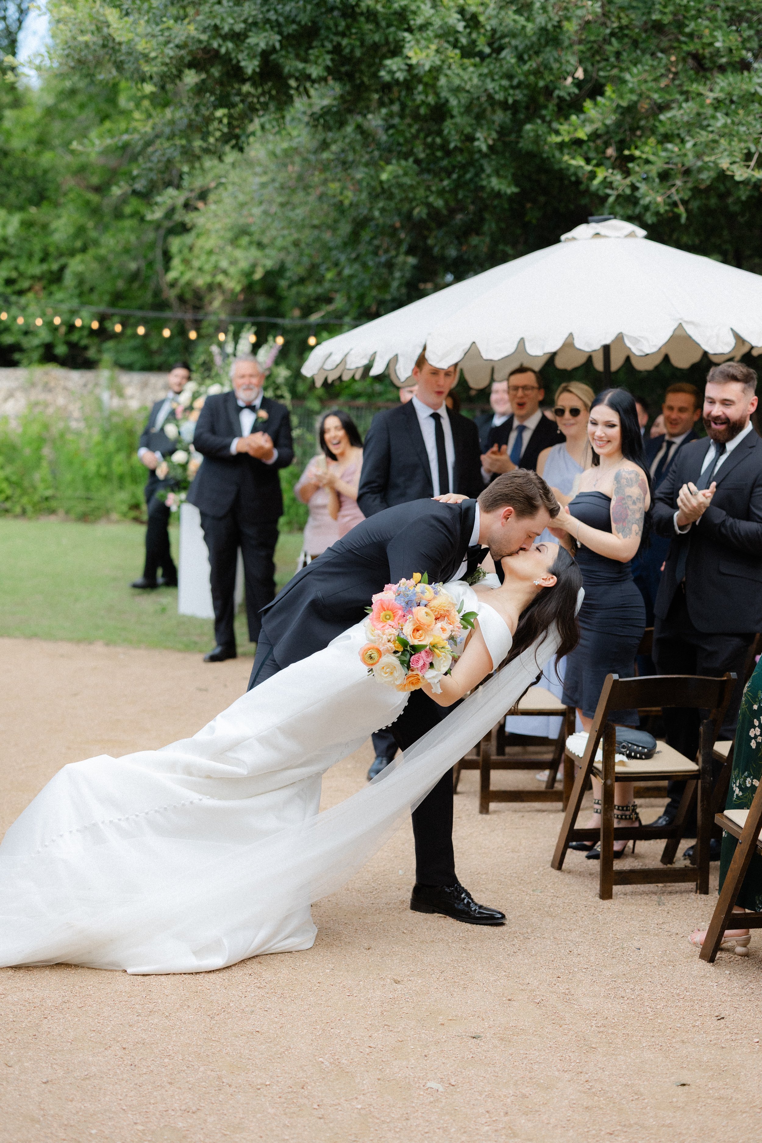 A wedding couple shares a kiss during their outdoor ceremony, with guests clapping and smiling in the background under a white umbrella.