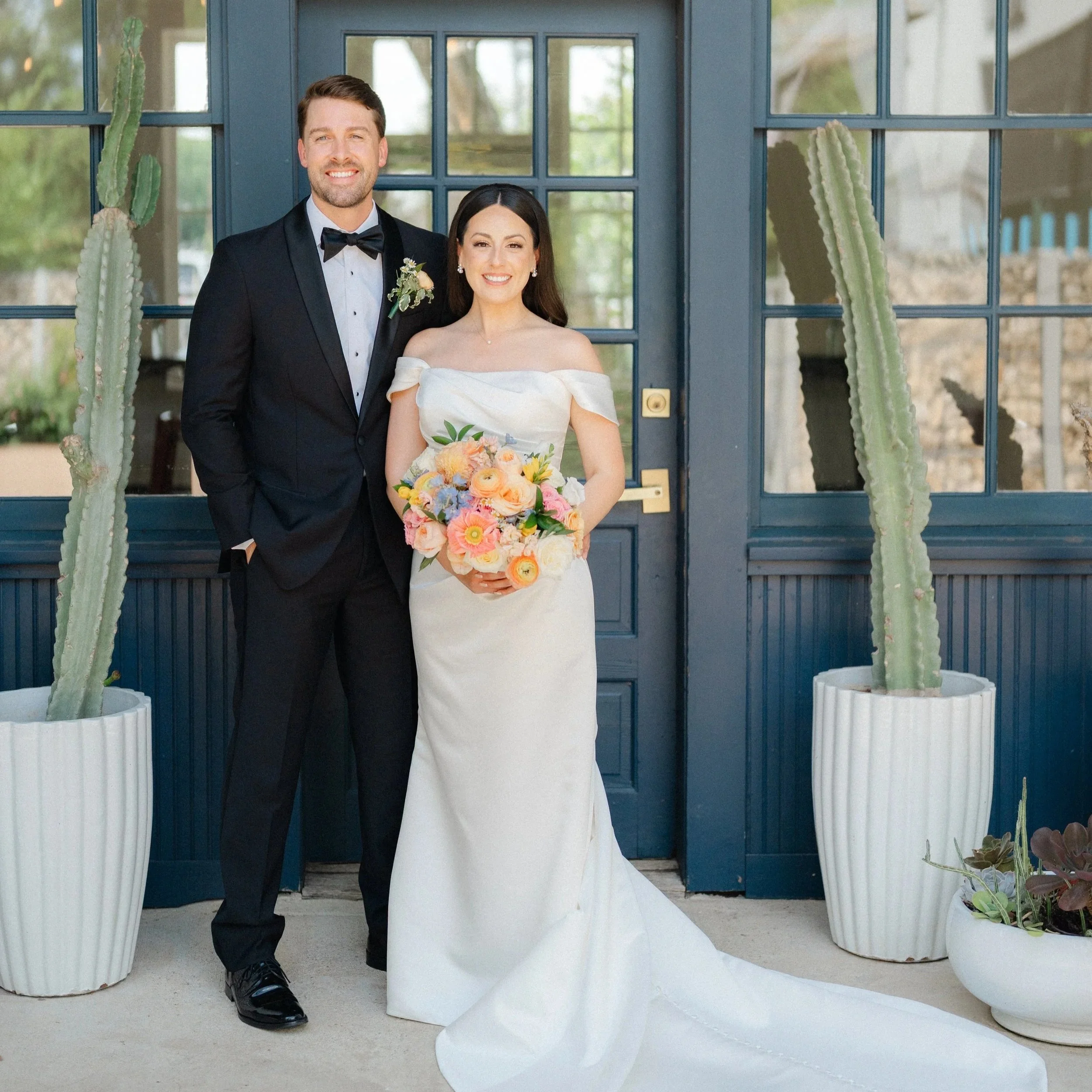 A bride and groom standing outside in front of a blue door with large potted cactus plants, smiling for a wedding photo. The bride is holding a colorful bouquet, wearing a white off-shoulder wedding gown. The groom is dressed in a black tuxedo with a bow tie.