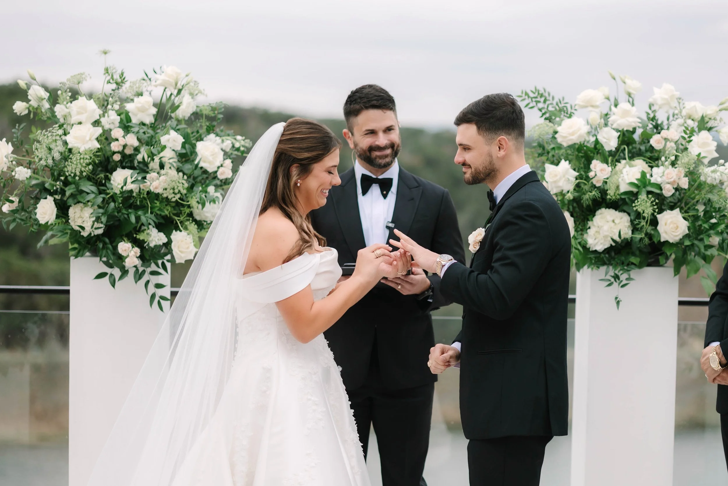 Bride and groom exchanging wedding rings at an outdoor wedding ceremony, with officiant and floral arrangements in the background.