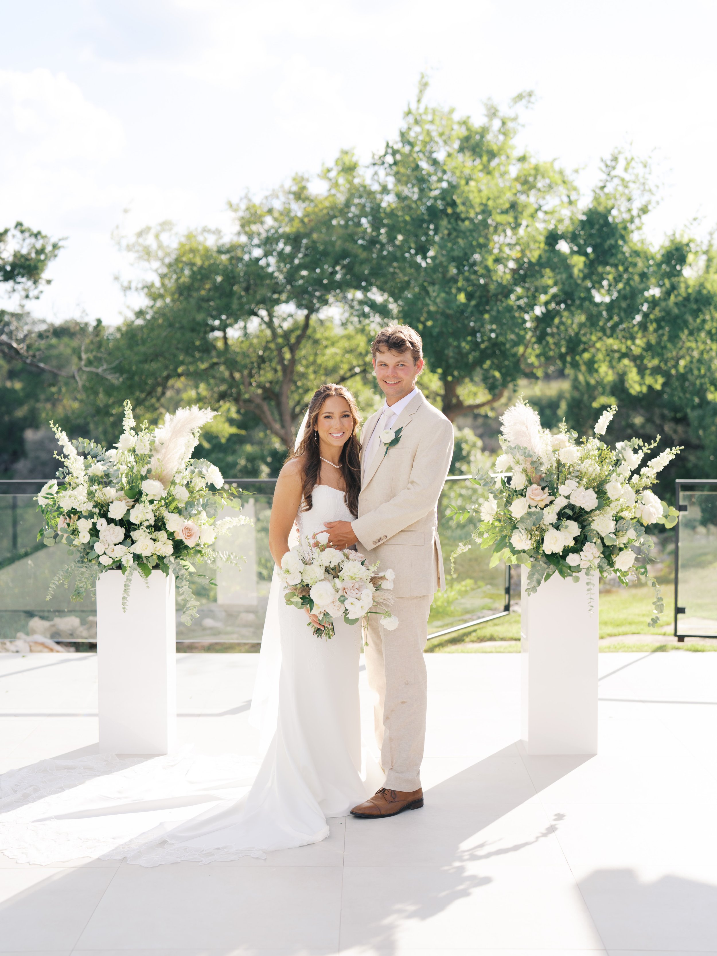 A bride and groom standing together outdoors on their wedding day, surrounded by large white floral arrangements, with a green tree and blue sky in the background.